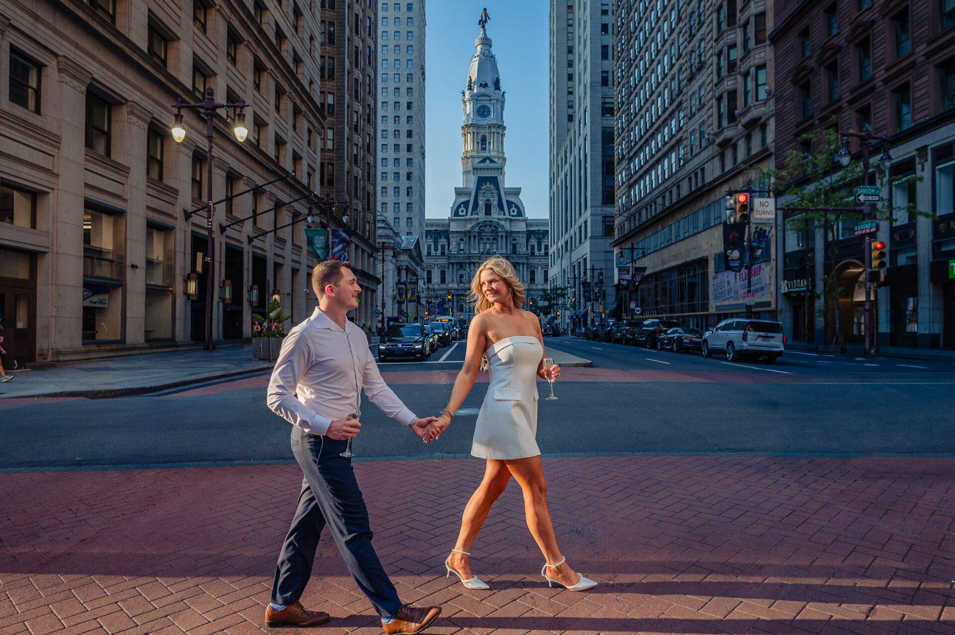 Engaged couple holding champagne glasses and walking across Broad Street with Philadelphia City Hall in background