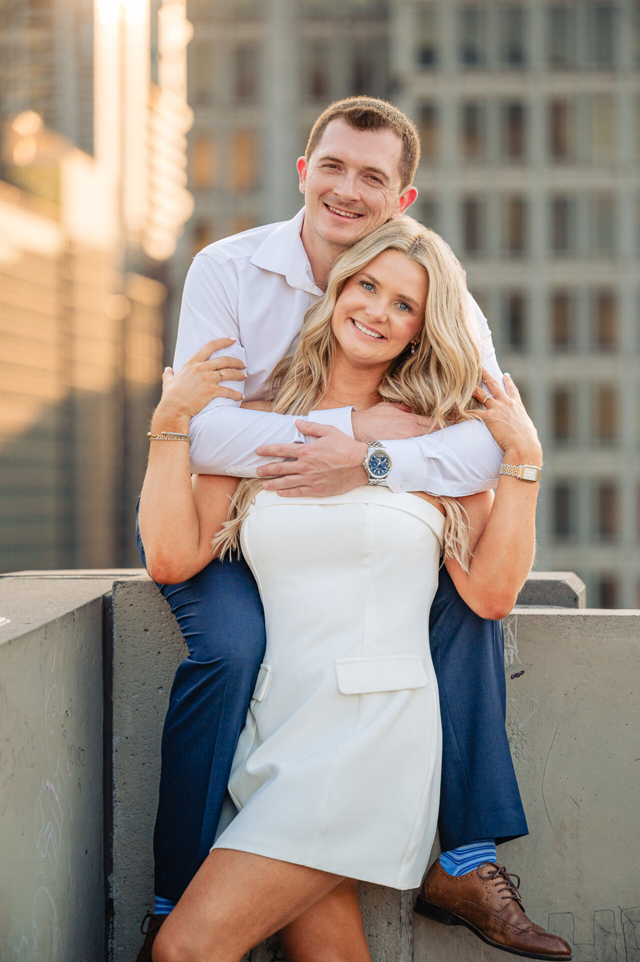Groom sitting behind bride on rooftop wall and hugging her tightly around shoulders while smiling at camera