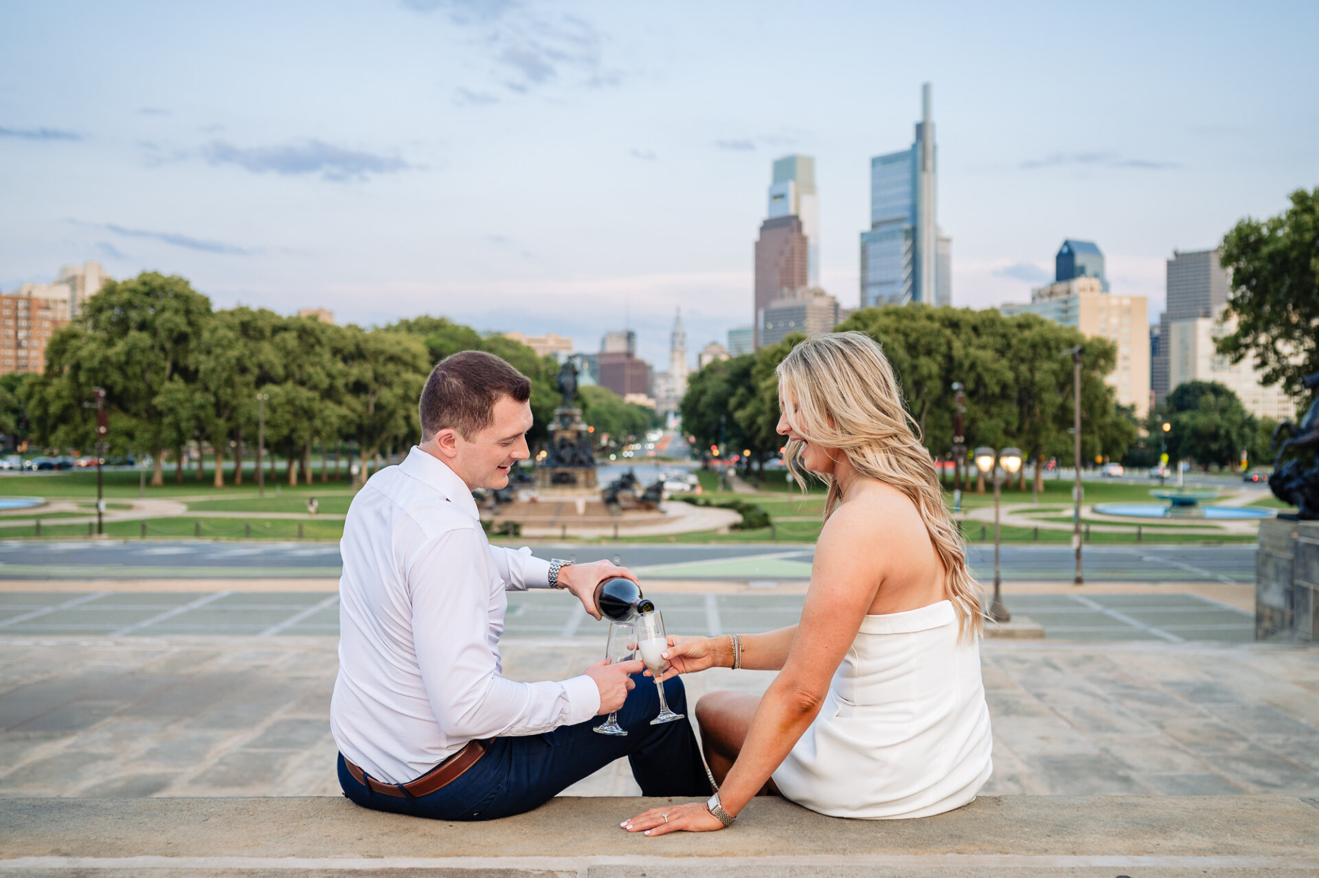 Groom pouring champagne into bride's glass while sitting outdoors with Philadelphia city view in background