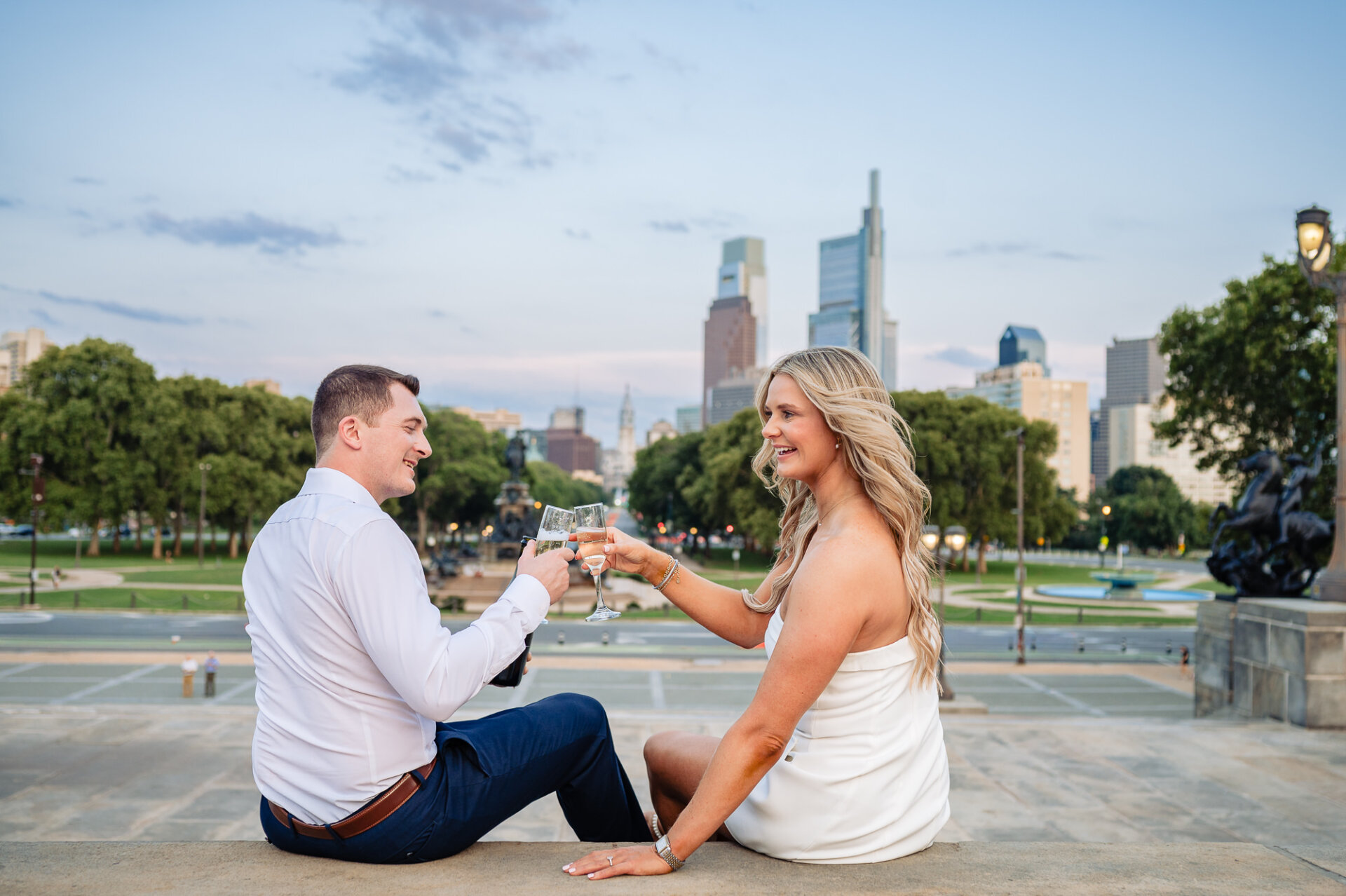 Engaged couple sitting on stone ledge and clinking champagne glasses with Philadelphia skyline in background