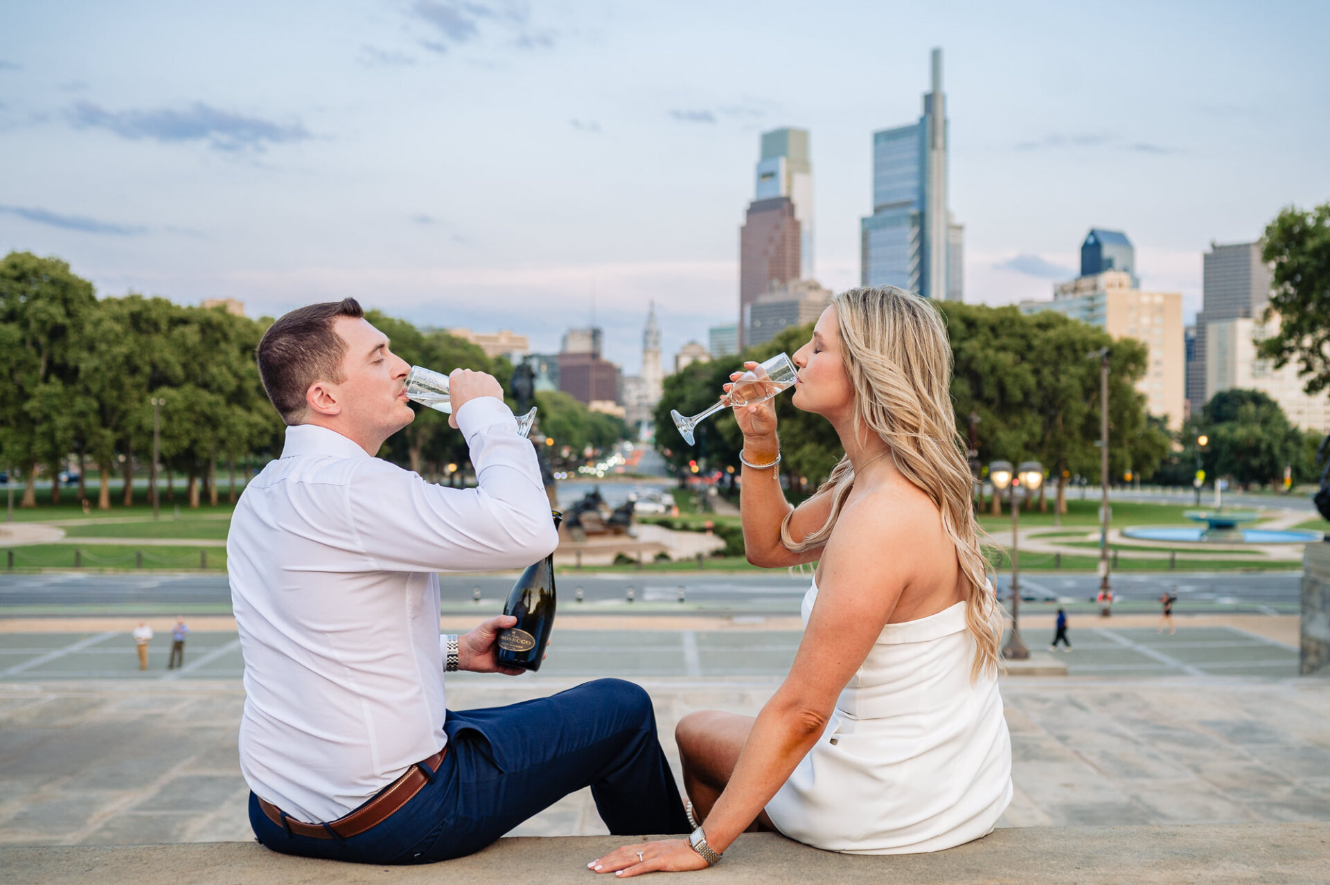 Side view of couple sitting on ledge and drinking prosecco with Philadelphia skyline framing background