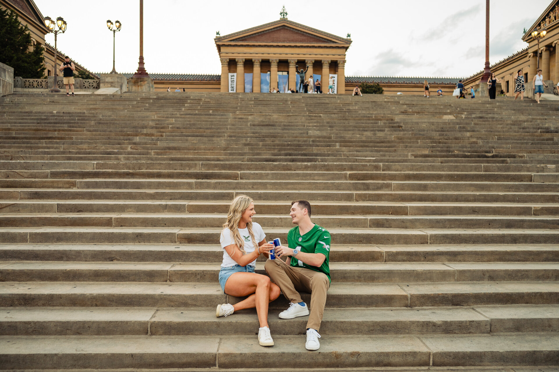 Engaged couple in Philadelphia Eagles gear sitting on Philadelphia Museum of Art steps and clinking beer cans