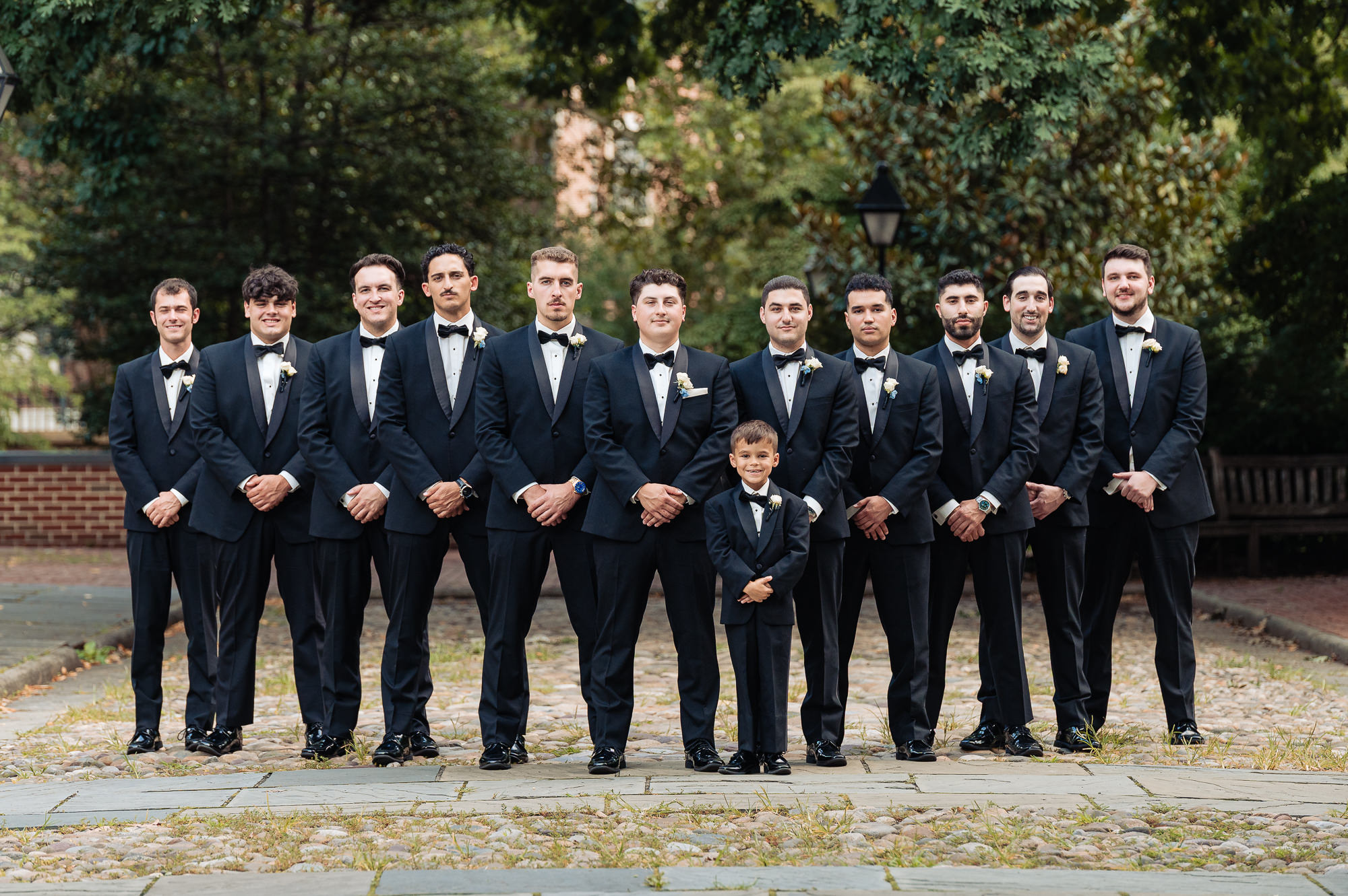 Groom and groomsmen in black tuxedos posing outdoors with a young ring bearer