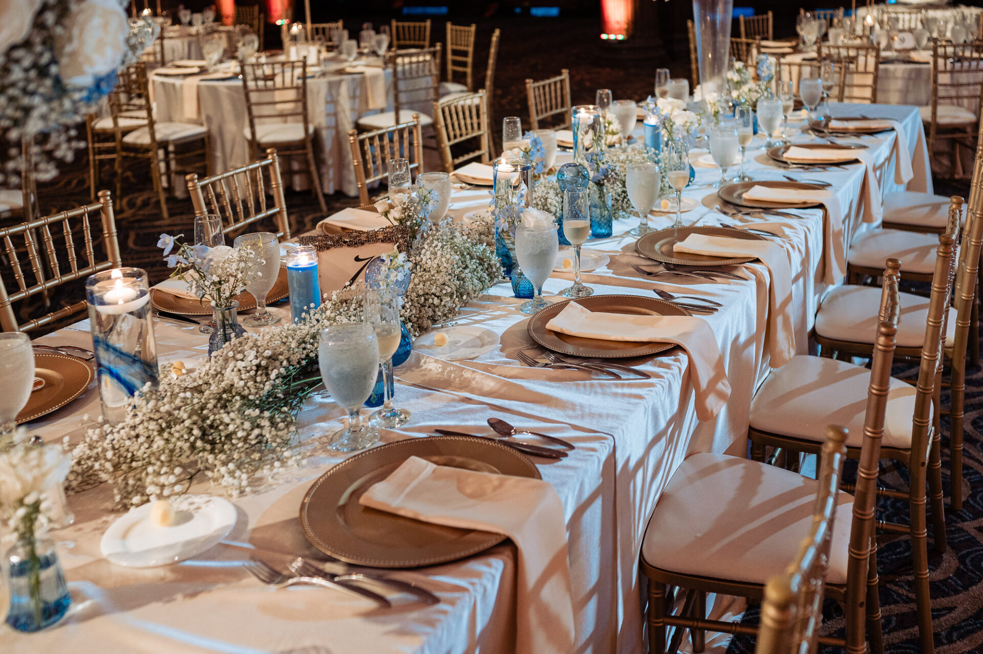 Wedding reception table decorated with baby’s breath, blue glass accents, candles, and gold place settings