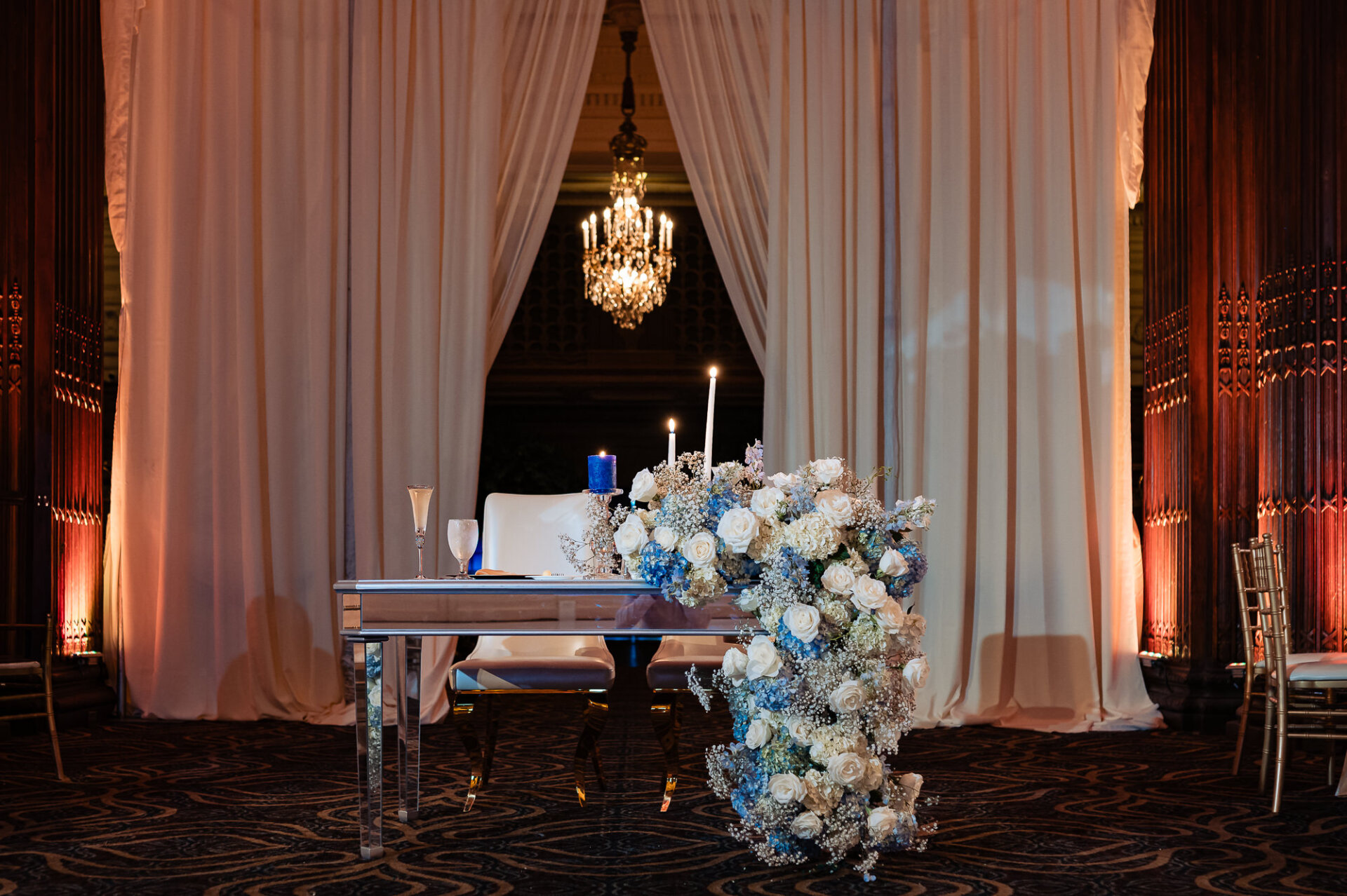 Sweetheart table decorated with cascading blue and white flowers, candles, and white draping