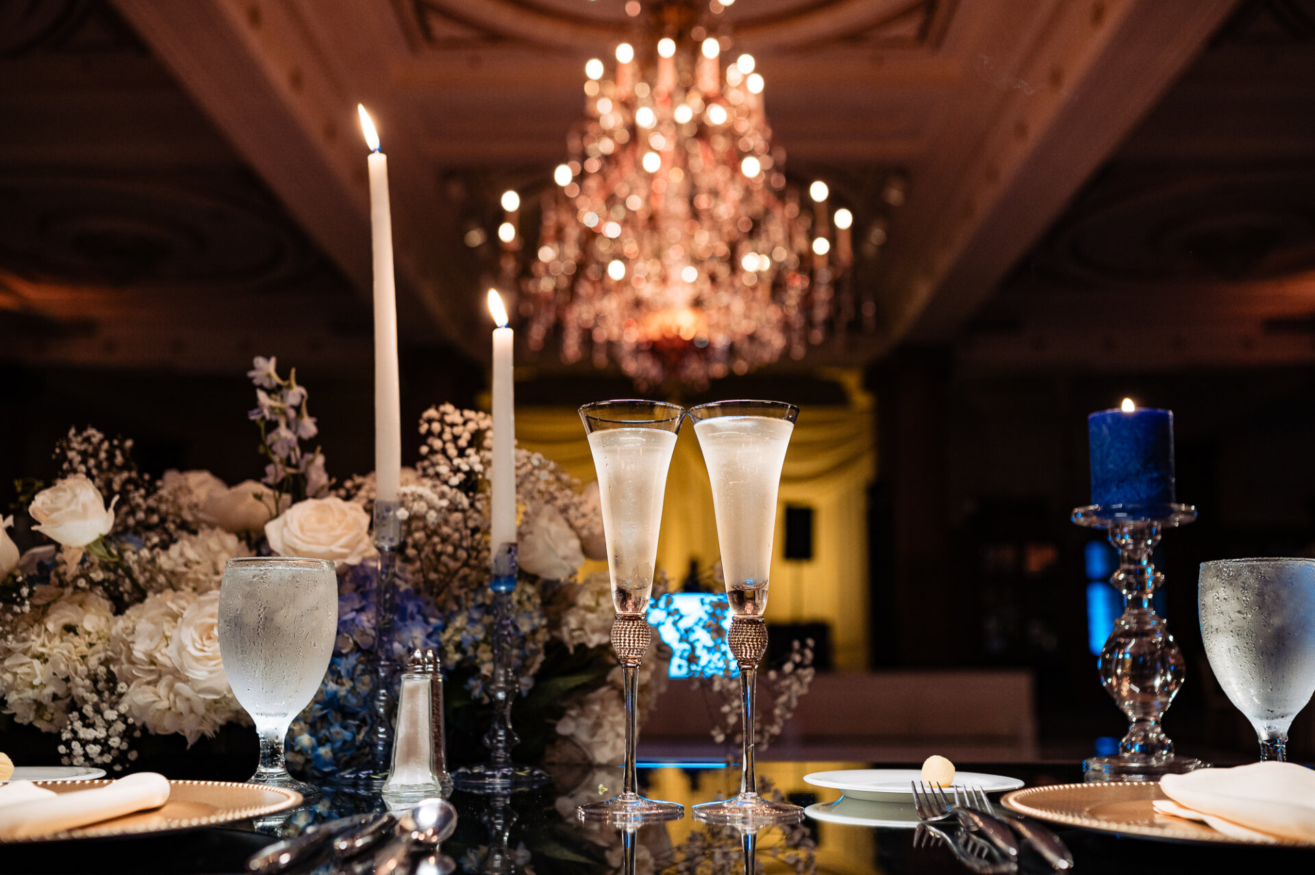 Wedding champagne glasses surrounded by candles and white florals beneath a sparkling crystal chandelier