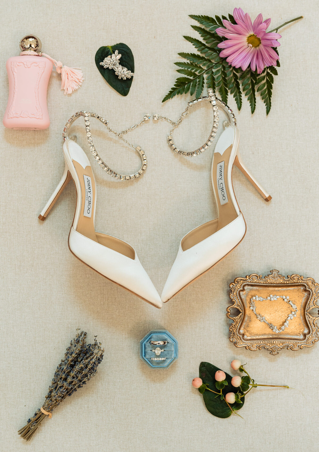 Bridal details flat lay with white Jimmy Choo heels, perfume, jewelry, wedding rings, and flowers