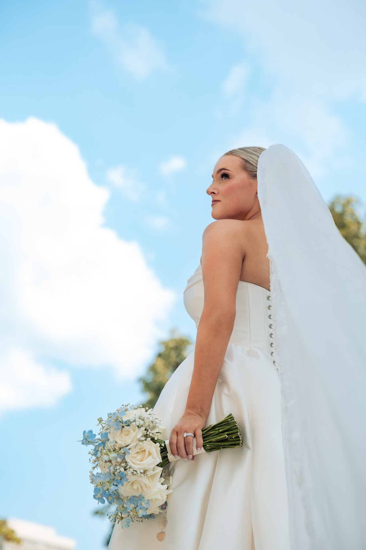 Low-angle bridal portrait showcasing a cathedral veil, wedding bouquet, and bright blue sky