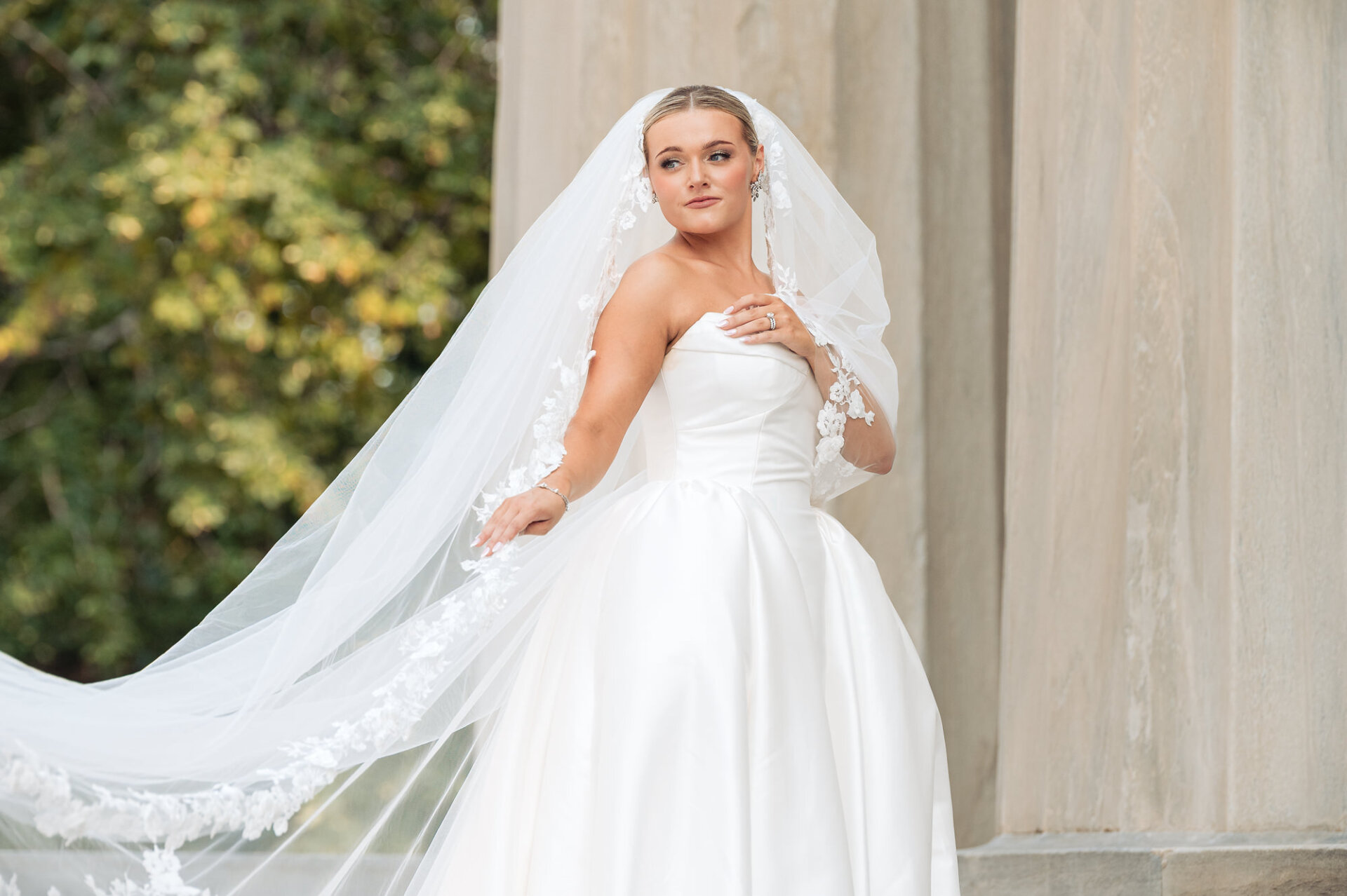 Bride posing beside a stone column with her long lace-trimmed veil flowing behind her