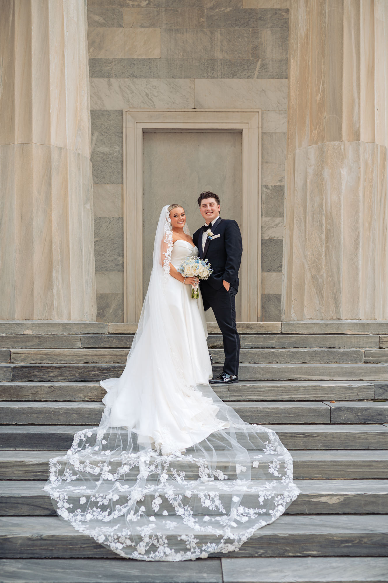 Bride and groom portrait on grand stone steps with the bride’s lace veil displayed behind them