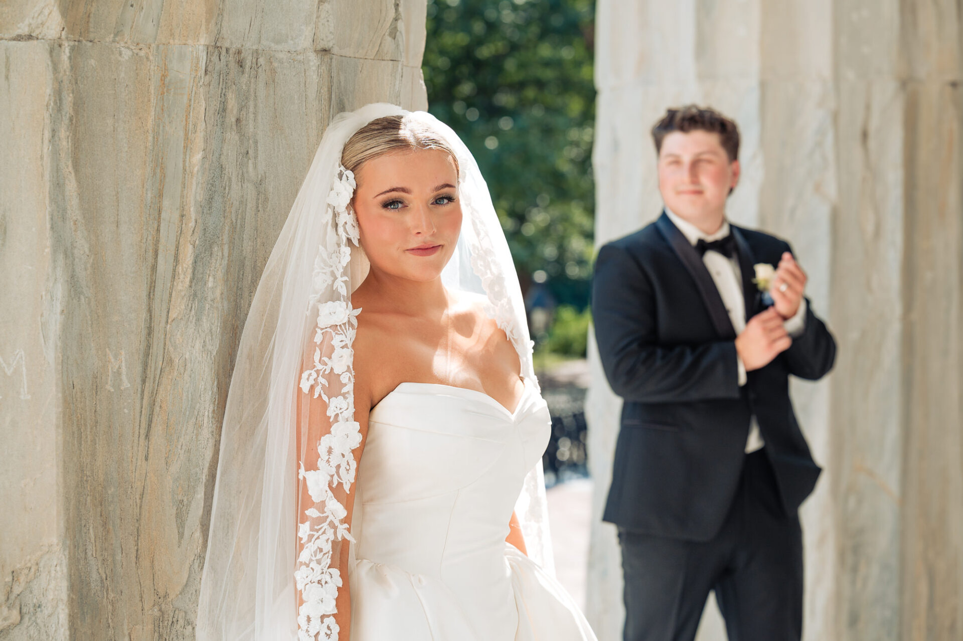 Bridal portrait framed by stone columns with the groom softly blurred in the background
