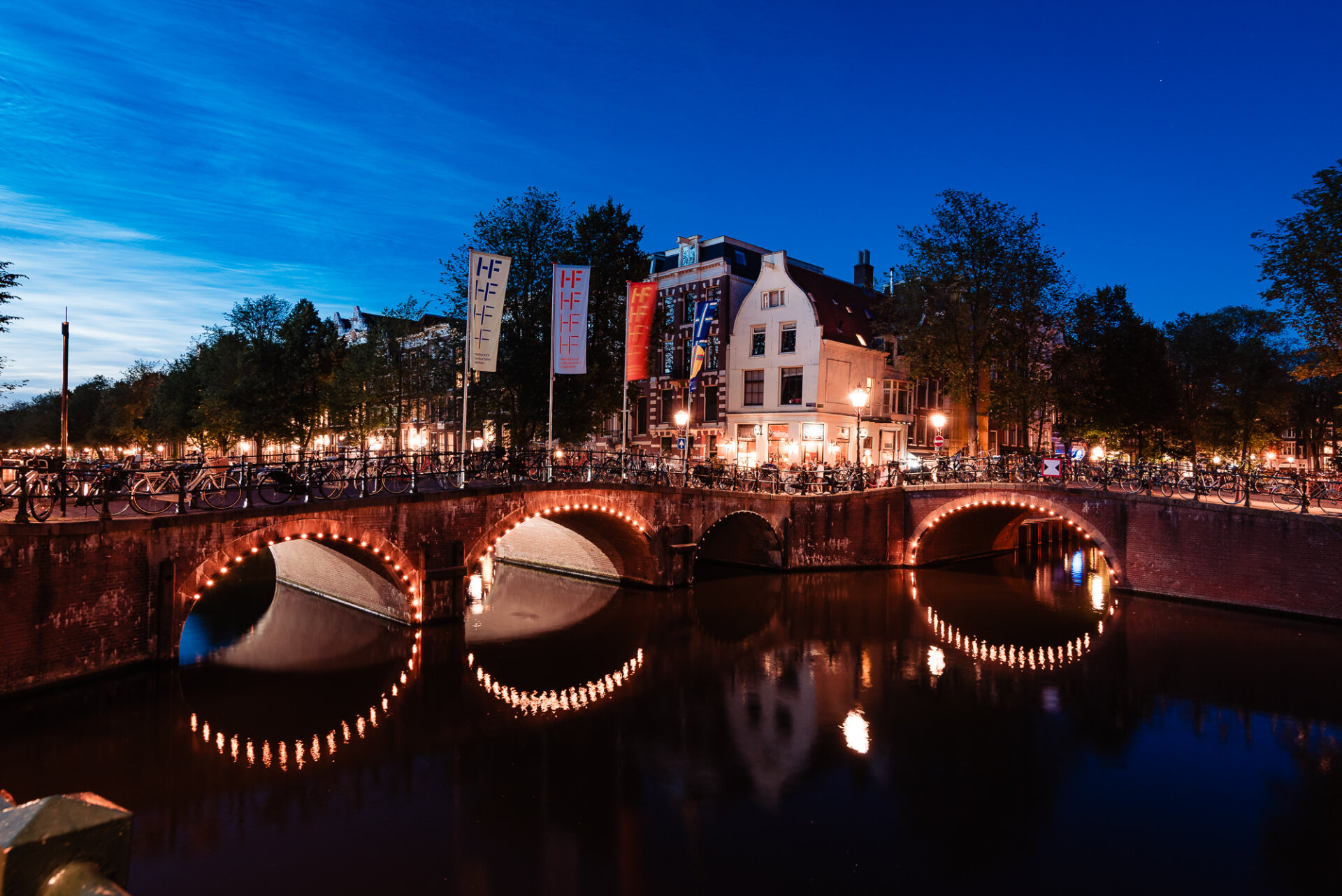 Illuminated canal bridges lined with bicycles reflecting across the water at blue hour in Amsterdam
