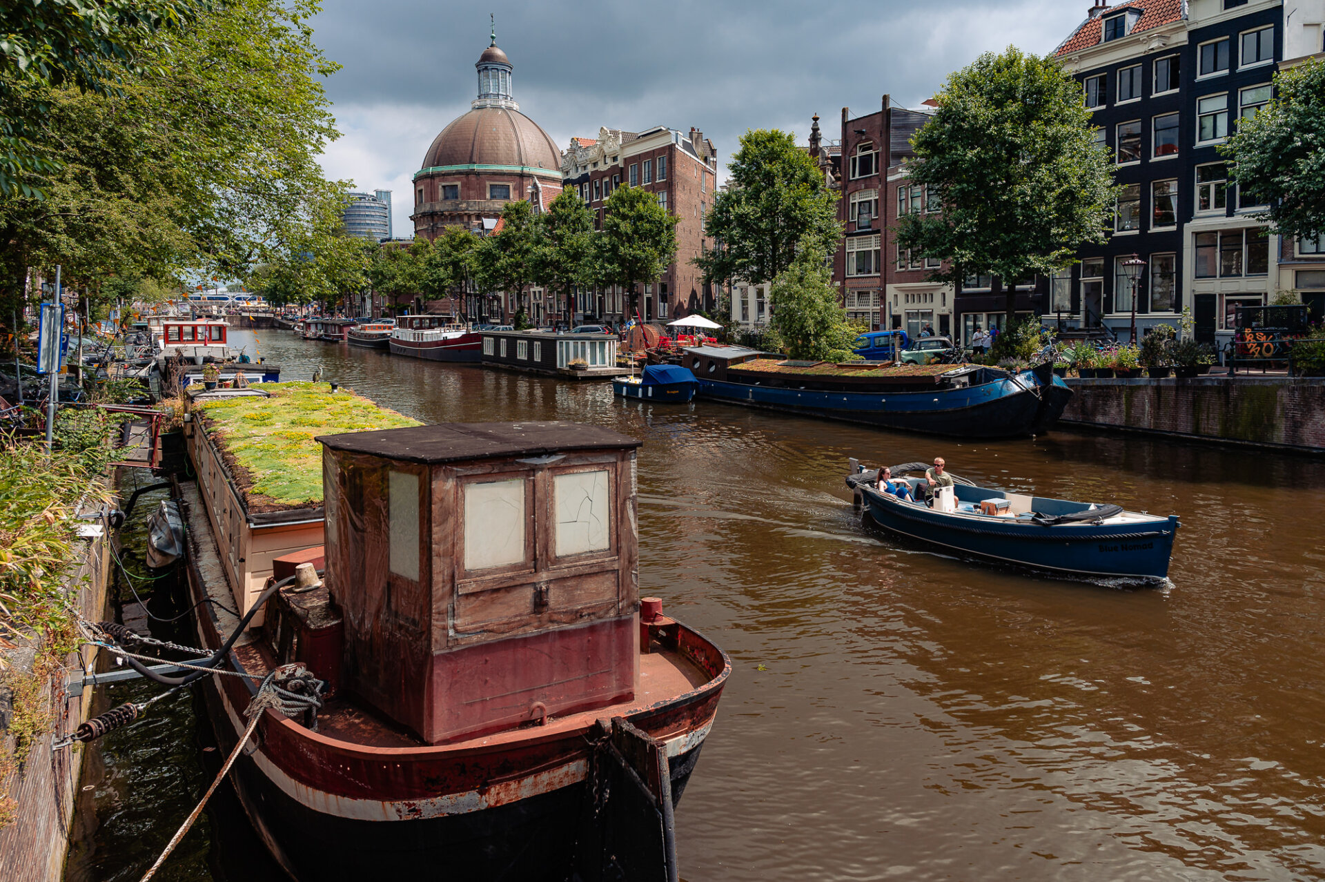 Houseboats and canal traffic passing historic Amsterdam buildings beneath a dramatic summer sky