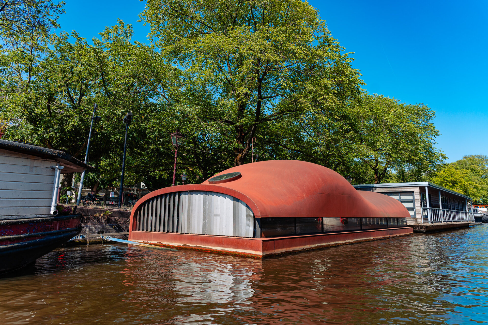 Distinctive modern houseboat with a curved red roof along a tree-lined Amsterdam canal