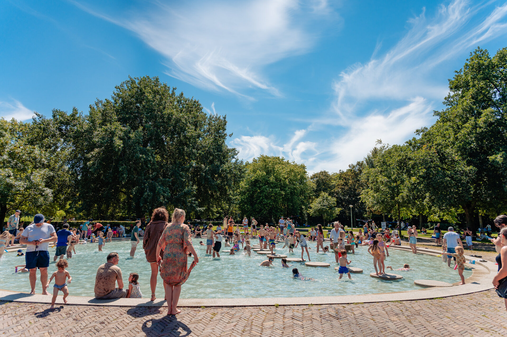 Families cooling off in a busy outdoor paddling pool surrounded by trees in Amsterdam