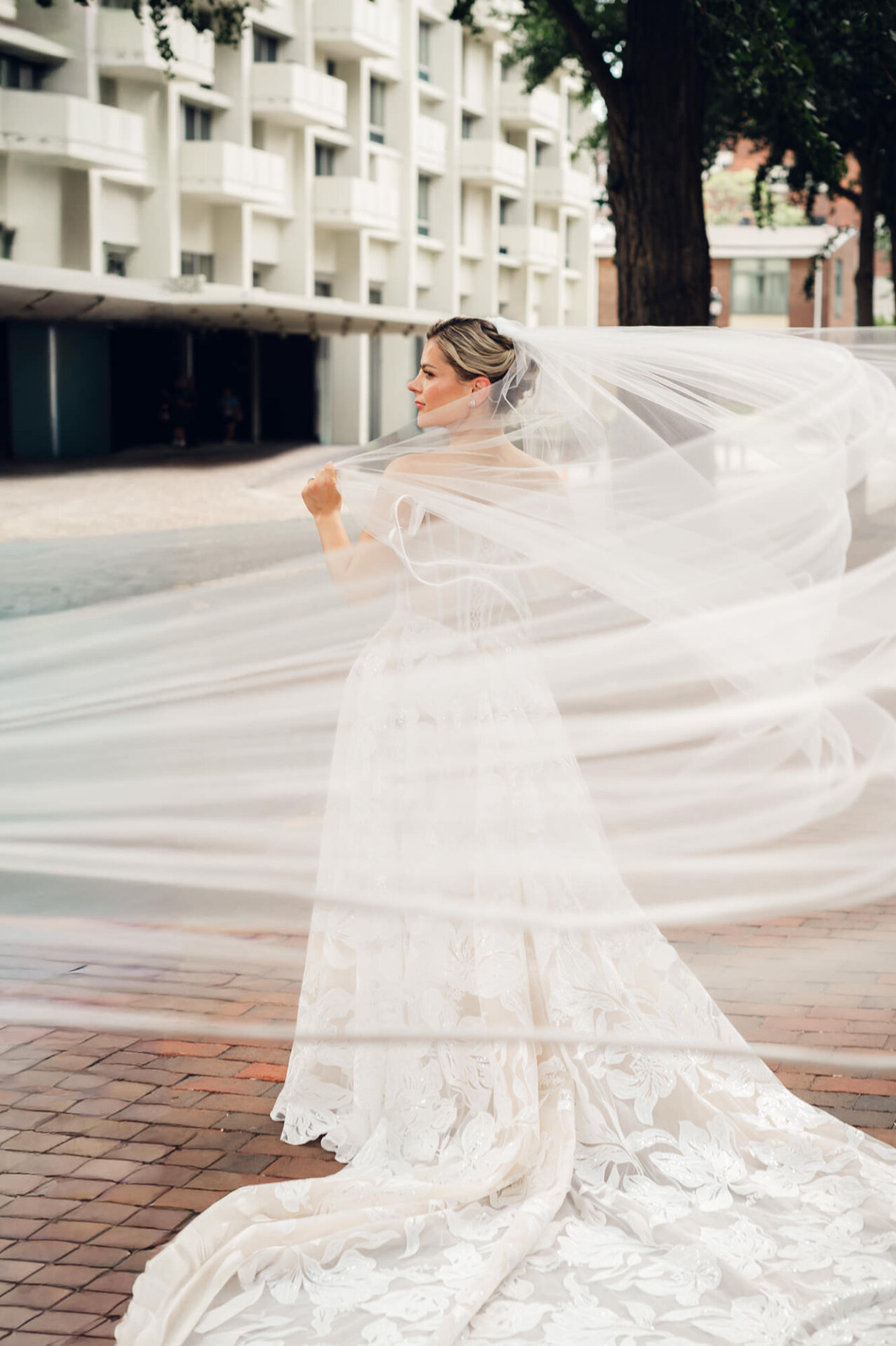 Bride poses outdoors in Philadelphia as her long veil flows around her floral lace wedding gown.