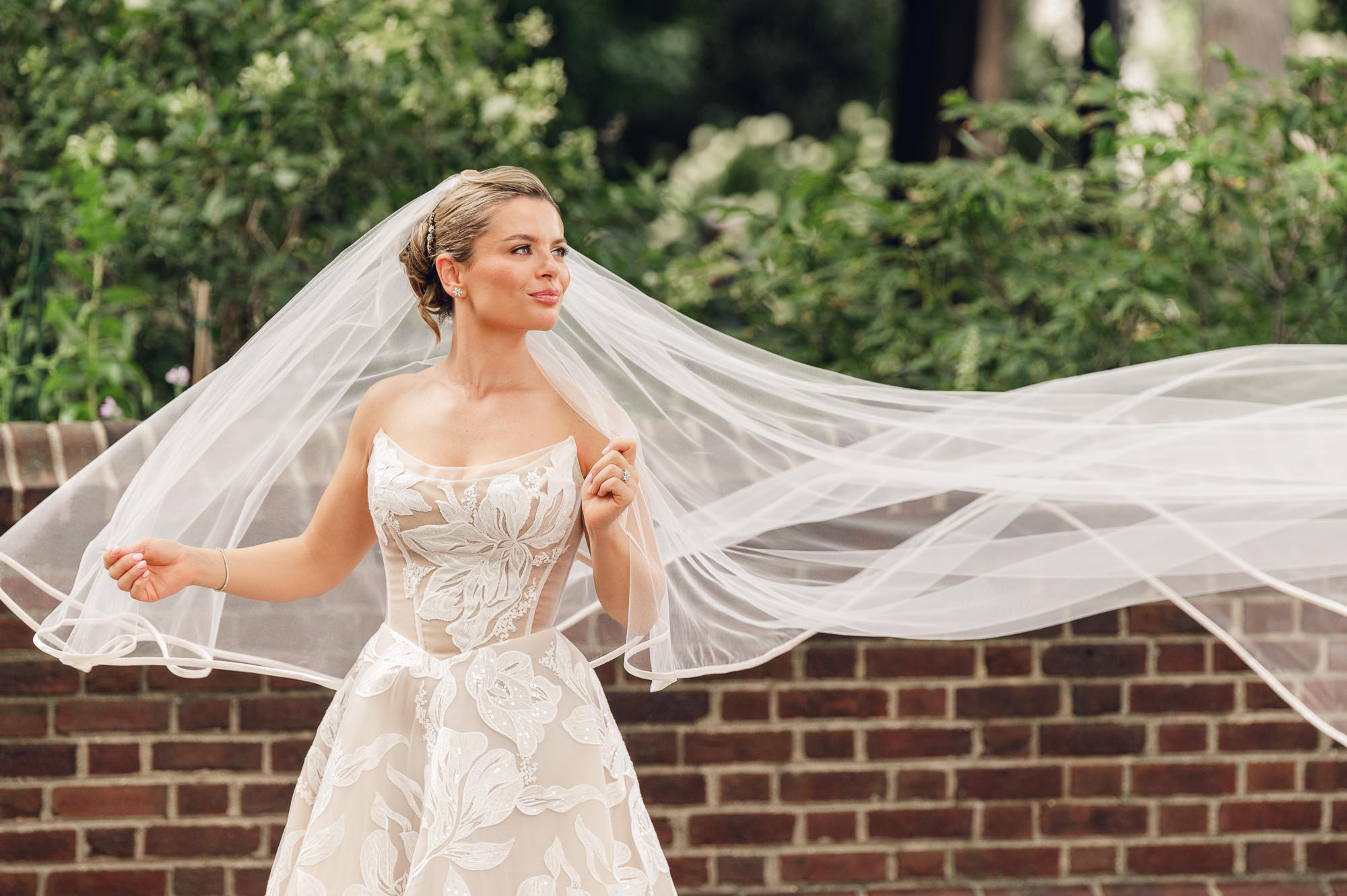 Bride poses outdoors in a floral lace gown as her long veil flows beside a brick wall.