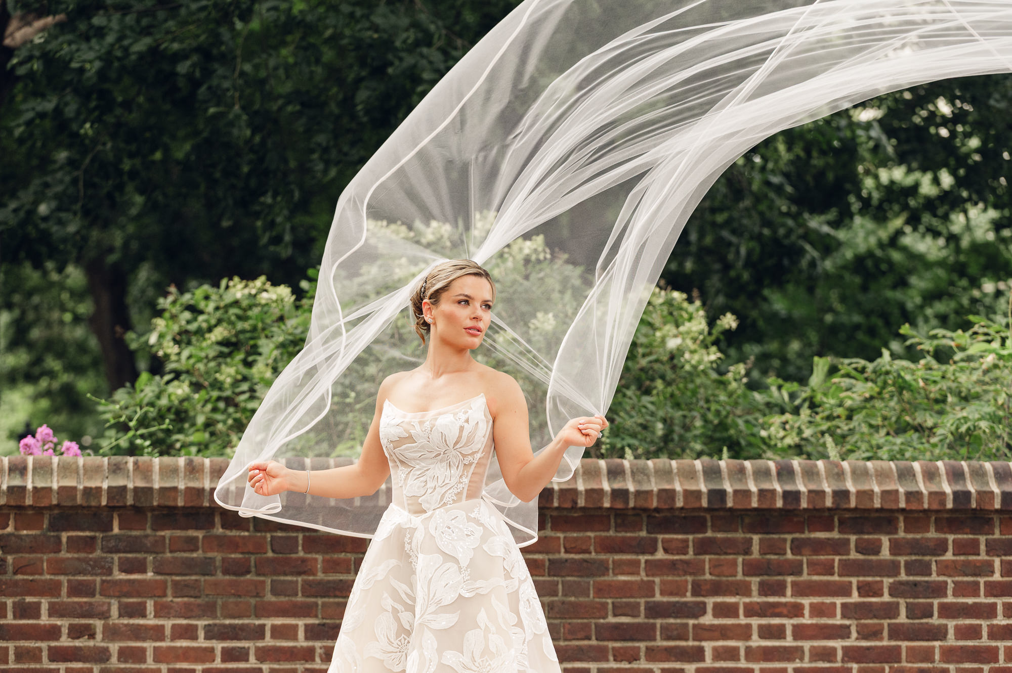 Bride holds her flowing veil for an outdoor wedding portrait beside a brick wall in Philadelphia.