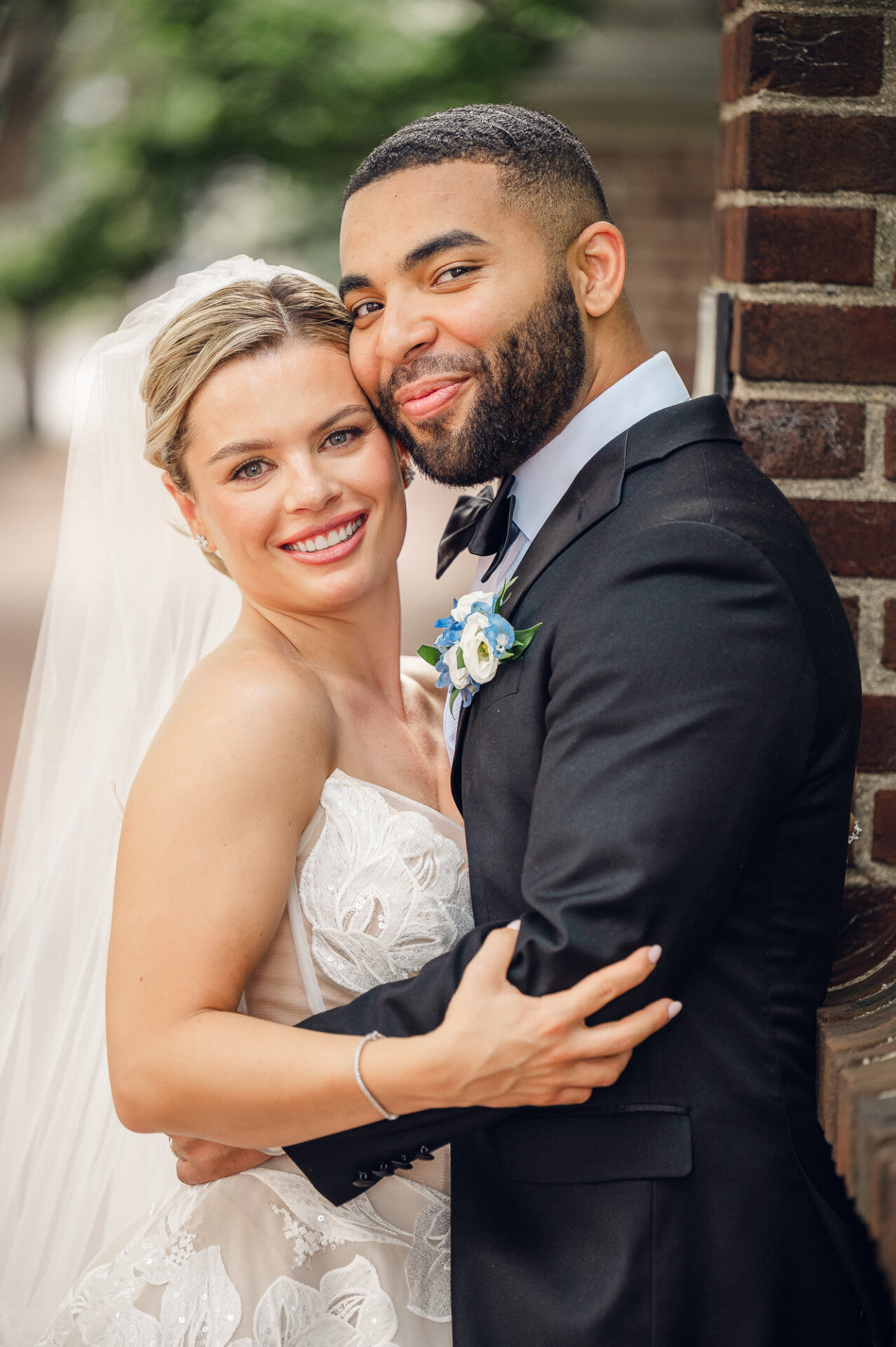 Close-up portrait of the bride and groom embracing by a brick wall, both smiling at the camera outdoors.