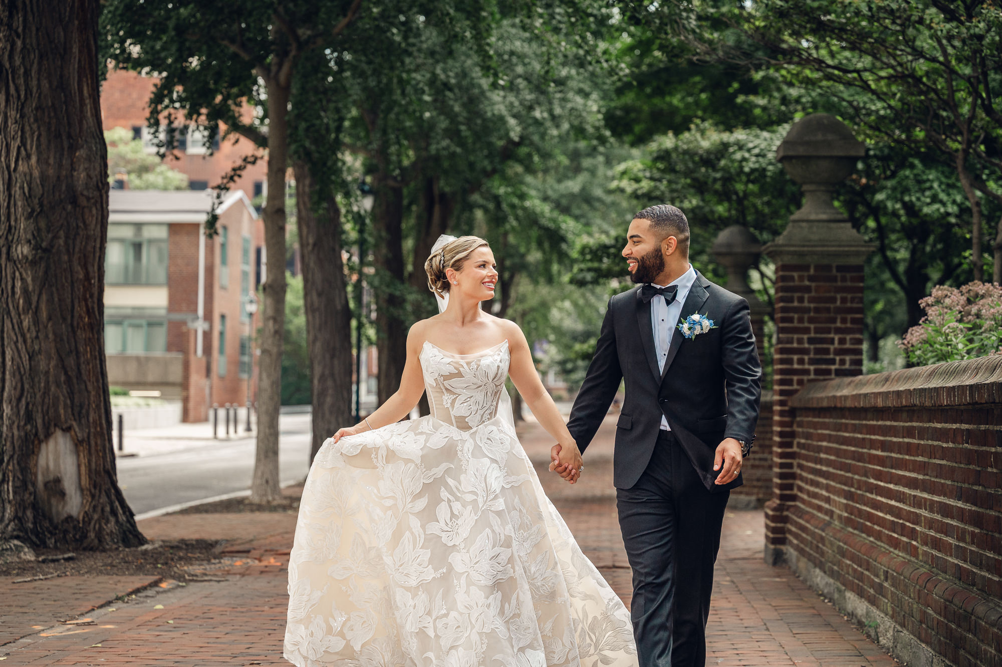 Bride and groom holding hands and smiling at each other while walking outdoors; bride wears a strapless floral lace ballgown and veil, groom wears a black tuxedo.