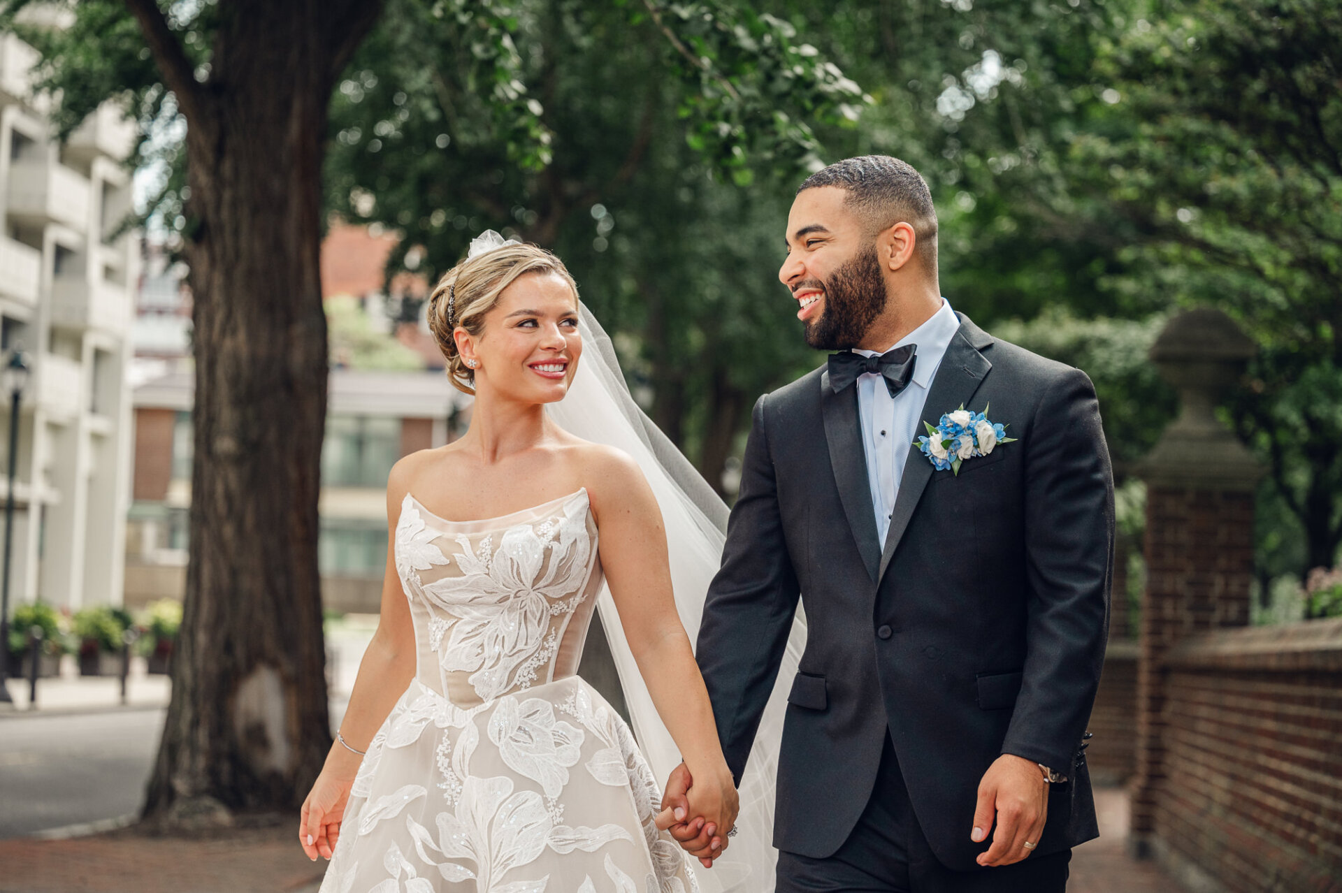 Groom smiling outdoors in a black tuxedo with a black bow tie, blue floral boutonniere, and polished dress shoes on a tree-lined walkway.