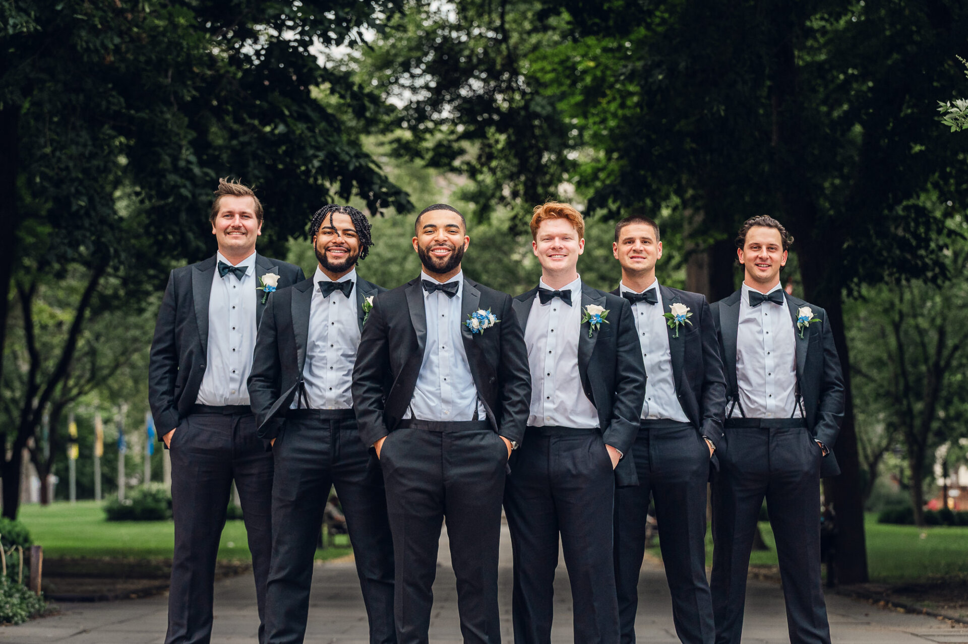 Groom and five groomsmen smiling in classic black tuxedos with black bow ties and blue and white boutonnieres outdoors on a paved walkway.