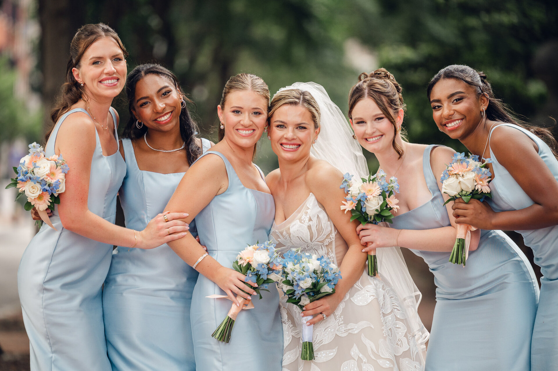 Bride smiling in her wedding dress surrounded by five bridesmaids in light blue square-neck satin dresses holding matching floral bouquets outdoors.