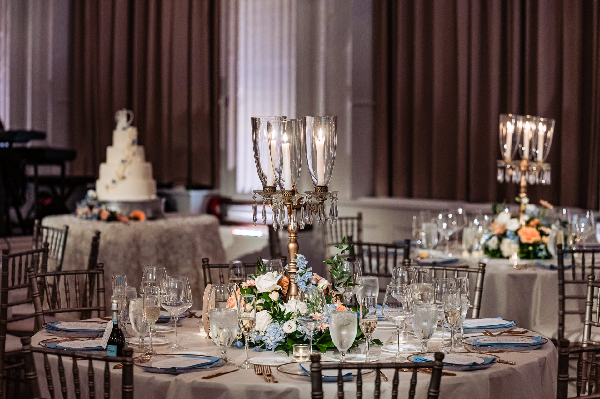 Tall gold candelabra centerpiece overflowing with peach roses, white roses, light blue delphiniums, and greenery on a set reception table.