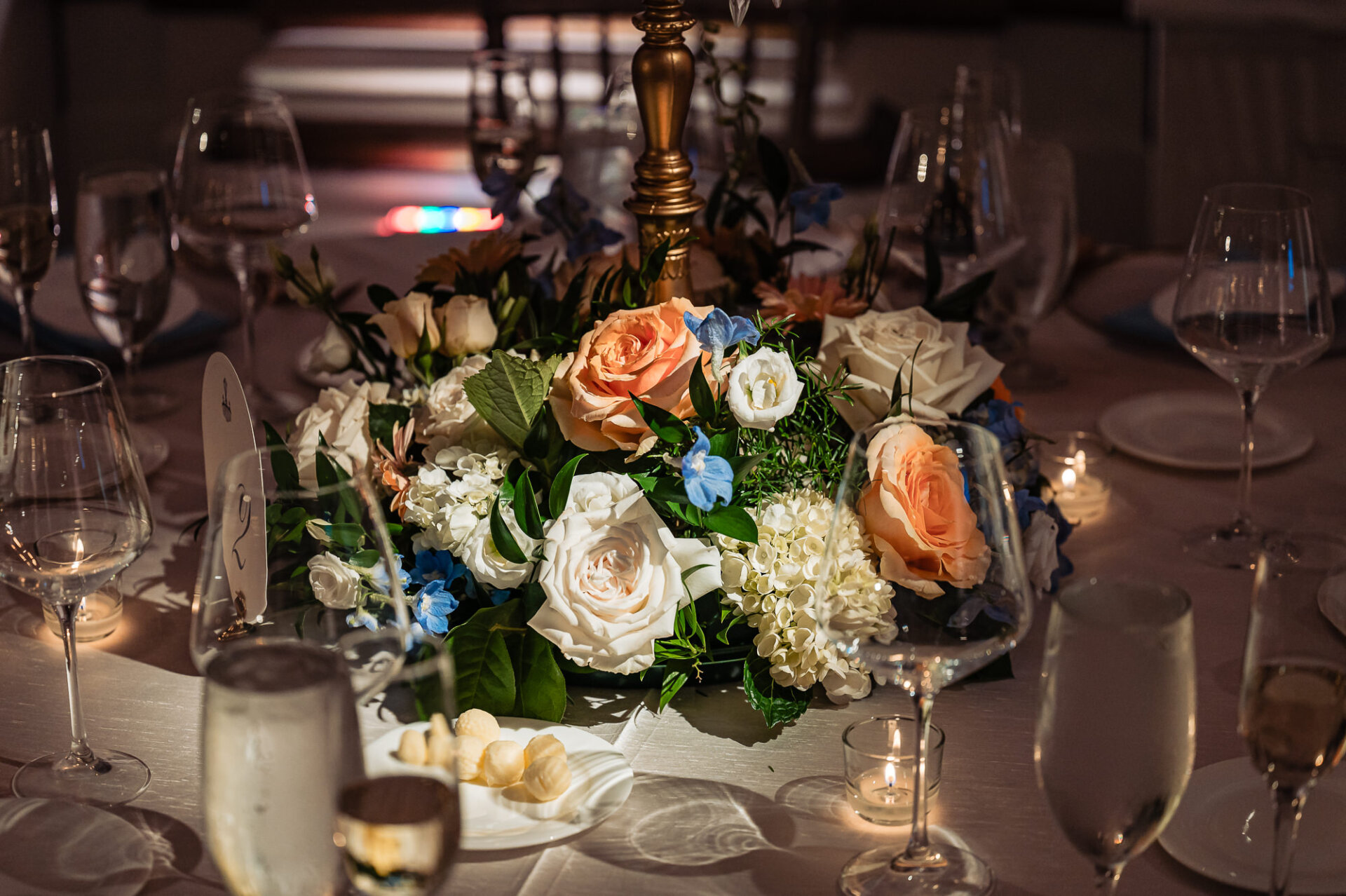 Low wedding reception table centerpiece featuring peach roses, white hydrangeas, white roses, and light blue delphiniums surrounded by wine glasses and candle votives.