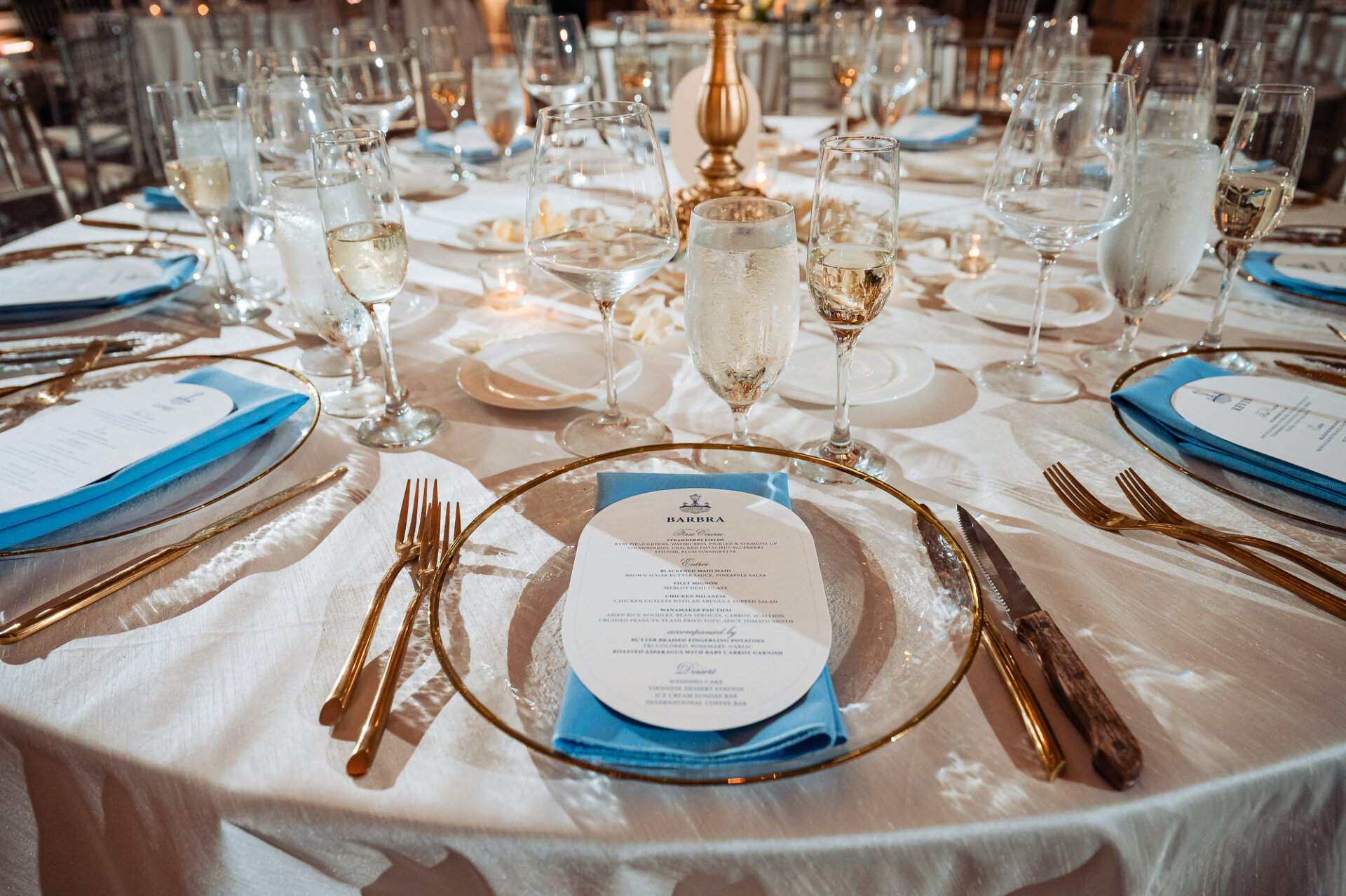 Wedding place setting featuring a clear glass charger plate with a gold rim, light blue folded napkin, printed dinner menu card, gold flatware, and wine glasses.