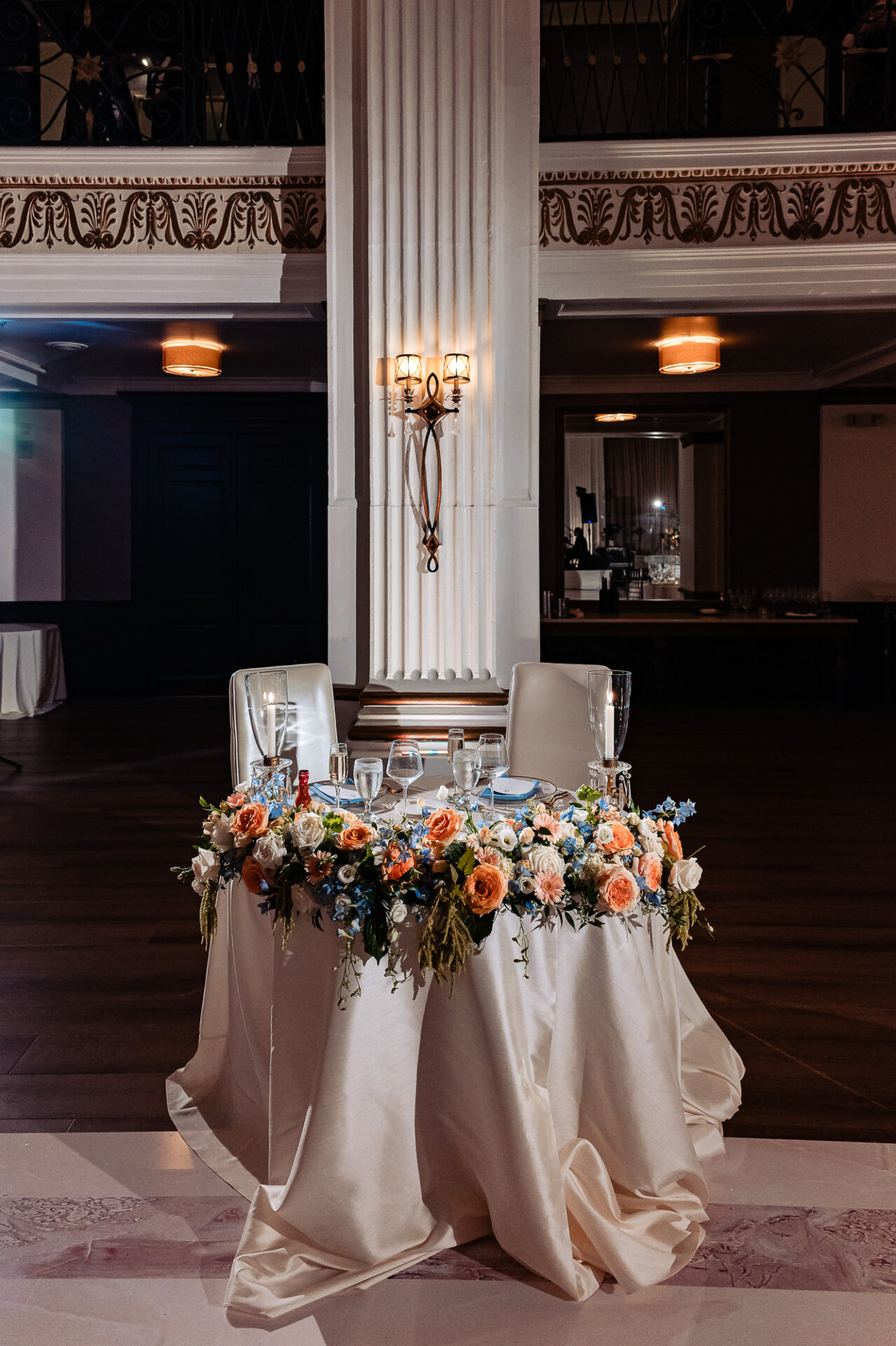 Elegant sweetheart table at the Ballroom at the Ben decorated with a lush cascading floral runner of peach, white, and blue flowers under a white column with wall sconces.