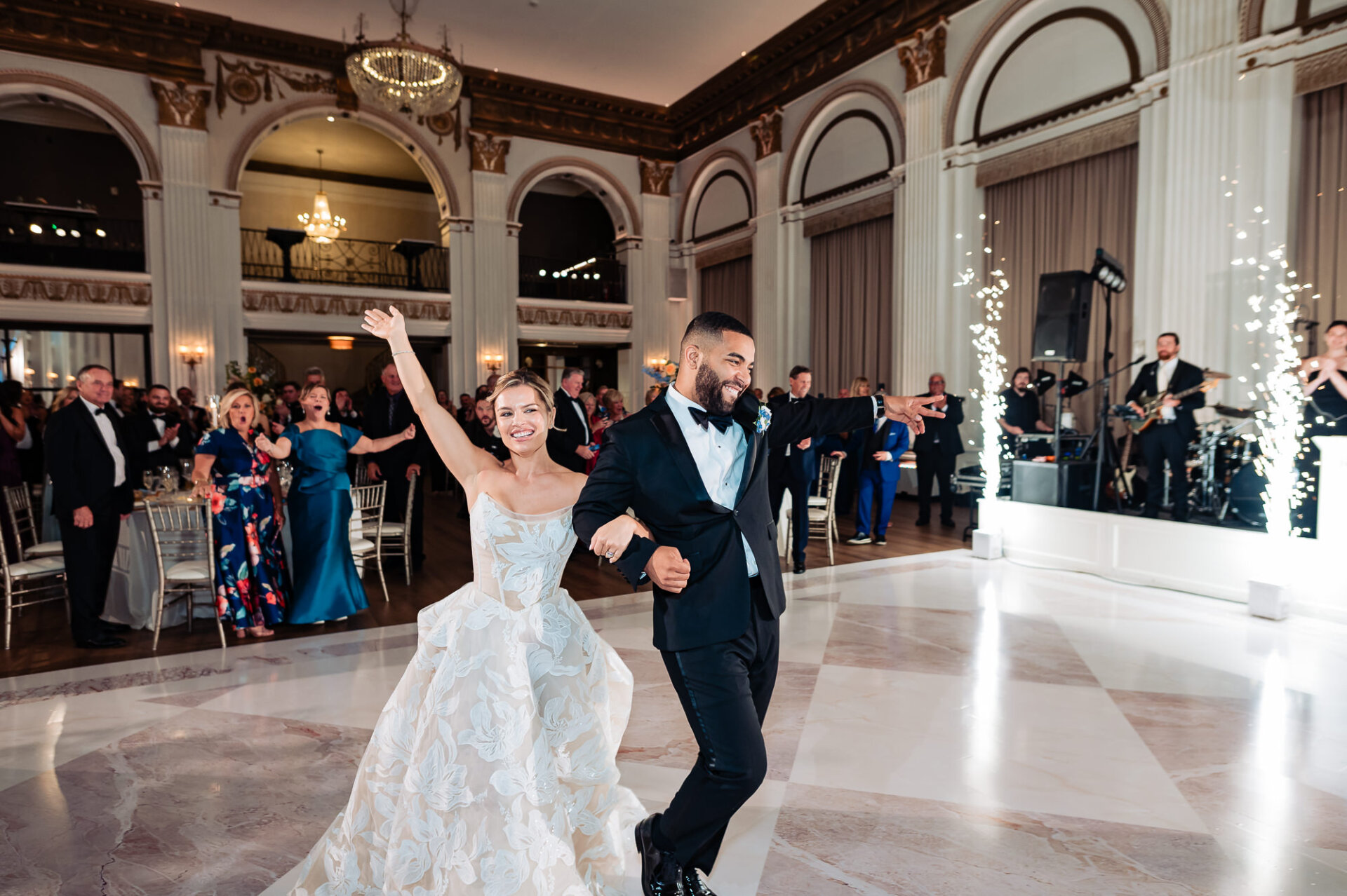 Newlyweds dancing into their wedding reception on a marble dance floor with arms outstretched, framed by indoor sparkler fountains and a live band stage.