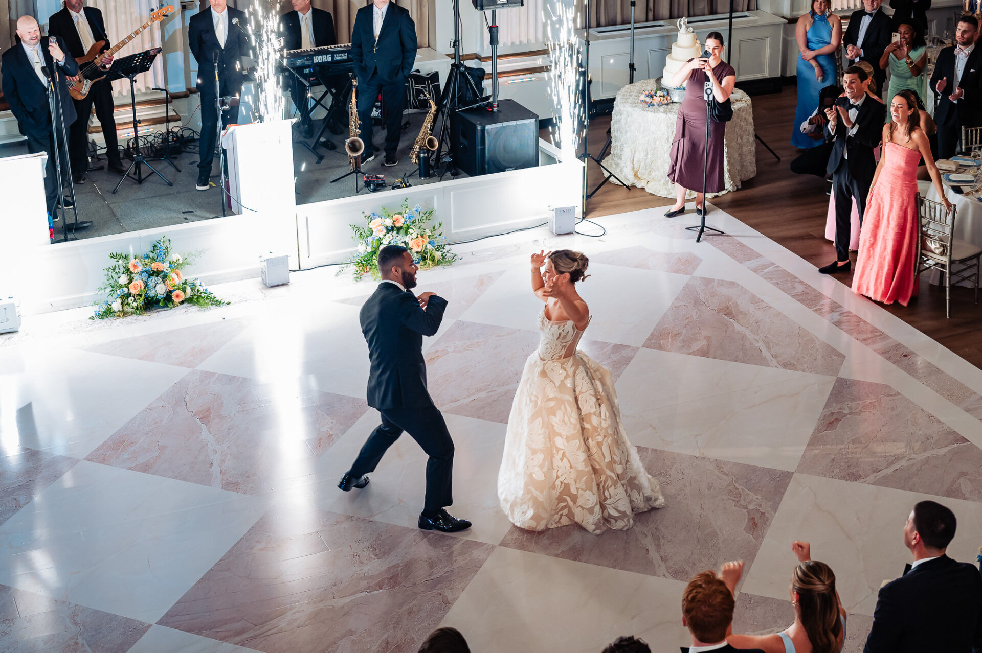 Overhead view of the bride and groom dancing together on a patterned marble dance floor flanked by indoor cold spark fountains and surrounded by cheering guests.