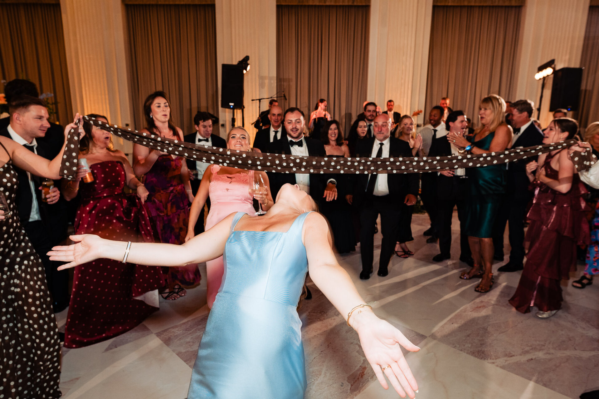 Wedding guest in a light blue dress doing the limbo under a polka dot scarf held by guests on a marble dance floor.