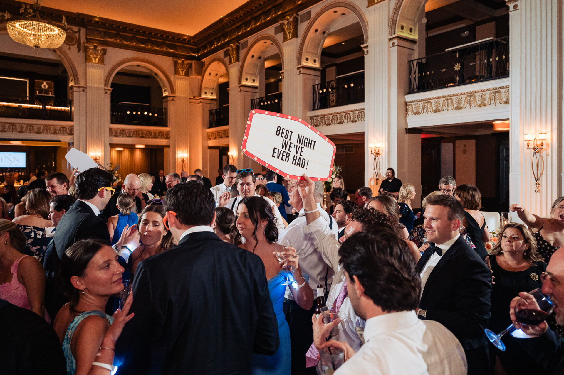Crowded dance floor at the Ballroom at the Ben during a wedding reception, with guests dancing and a man holding up a lighted marquee sign reading "BEST NIGHT WE'VE EVER HAD!"