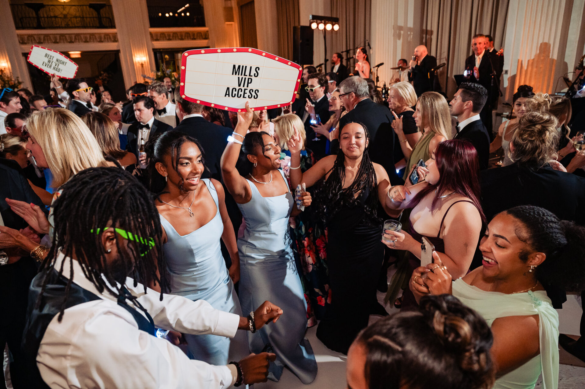 Guests dancing enthusiastically at a wedding reception, with one guest holding up a marquee sign that reads "MILLS VIP ACCESS".