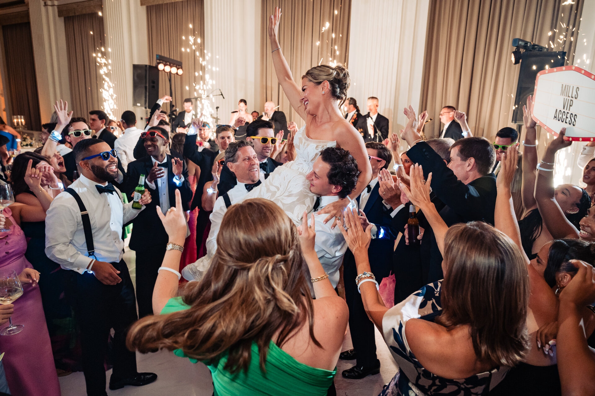 Bride being lifted into the air by wedding guests on a crowded dance floor while holding her arm up, surrounded by cheering guests and sparkler fountains in the background.