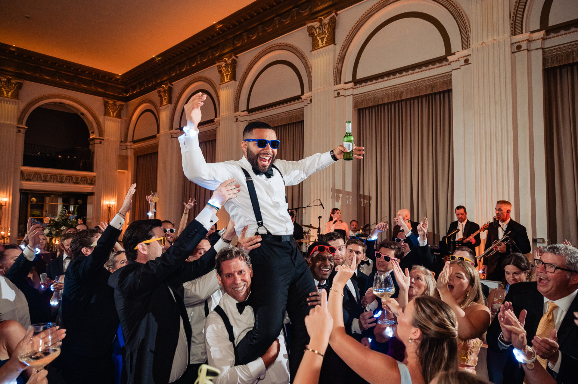 Groom wearing blue sunglasses hoisted on the shoulders of wedding guests while holding a beer on the dance floor during a reception.