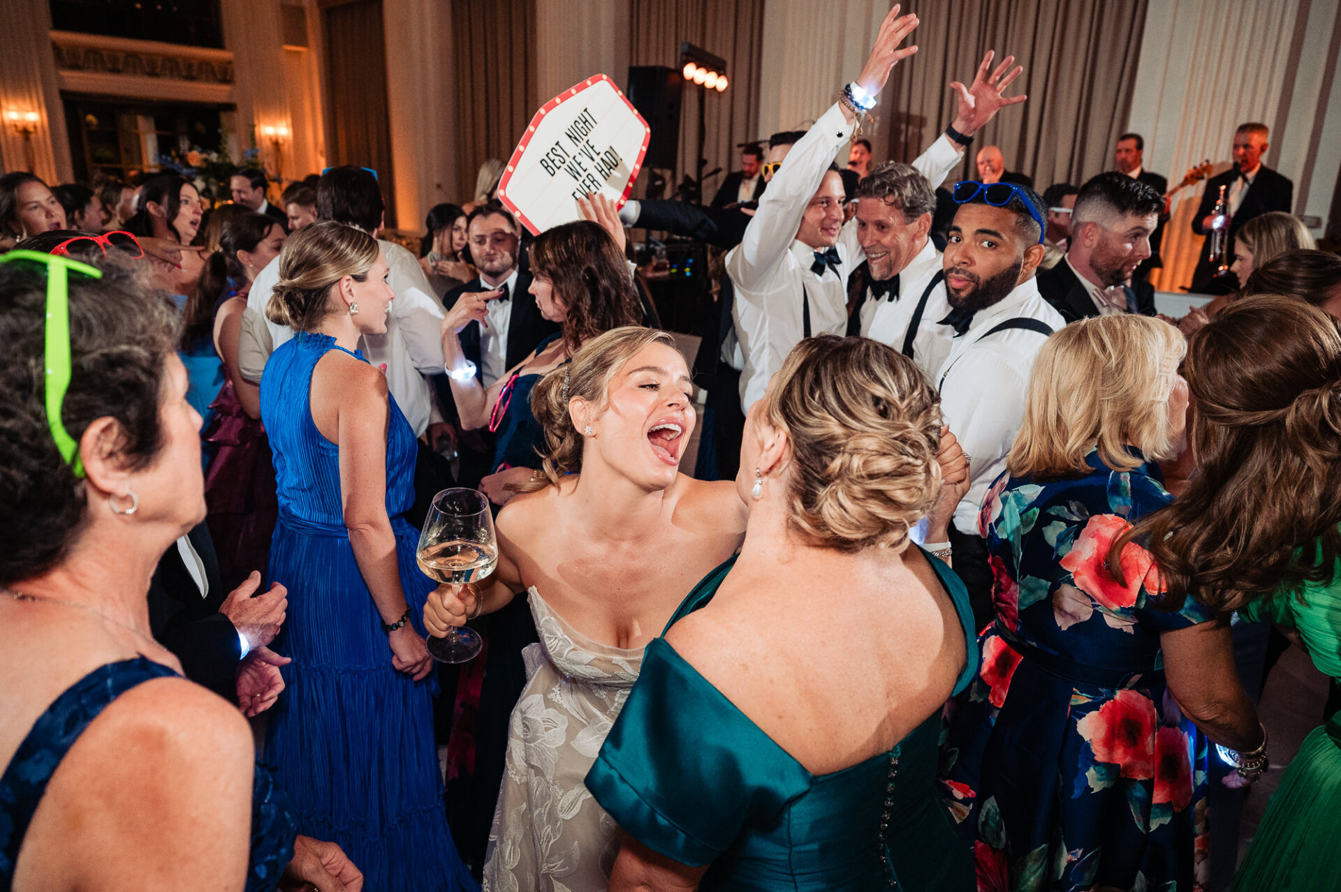 Bride in an embroidered strapless gown singing and dancing with a guest holding a wine glass on a packed dance floor.
