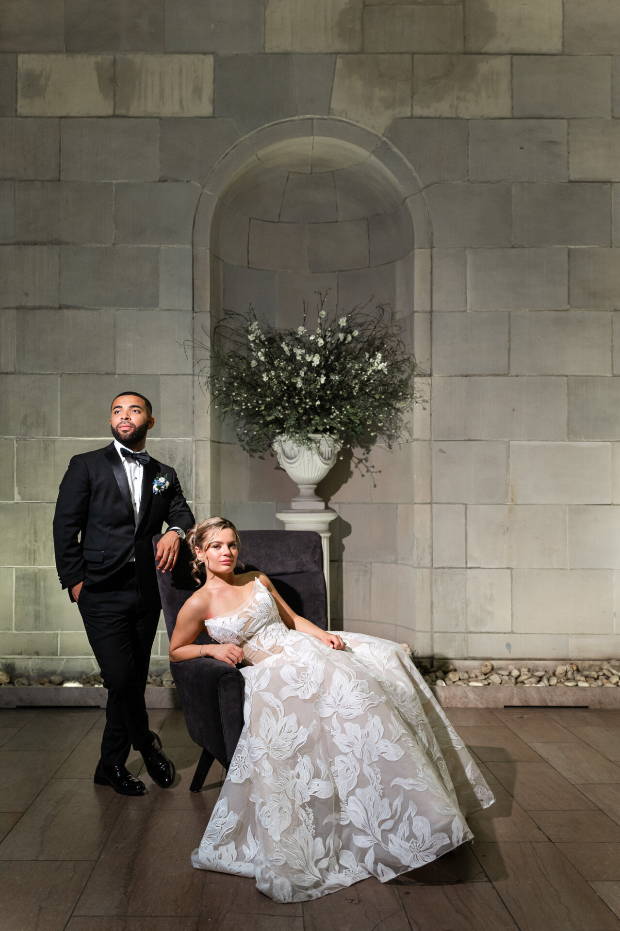 Formal portrait of the bride reclining in a dark armchair while the groom stands beside her leaning on the chair in a stone architectural alcove.