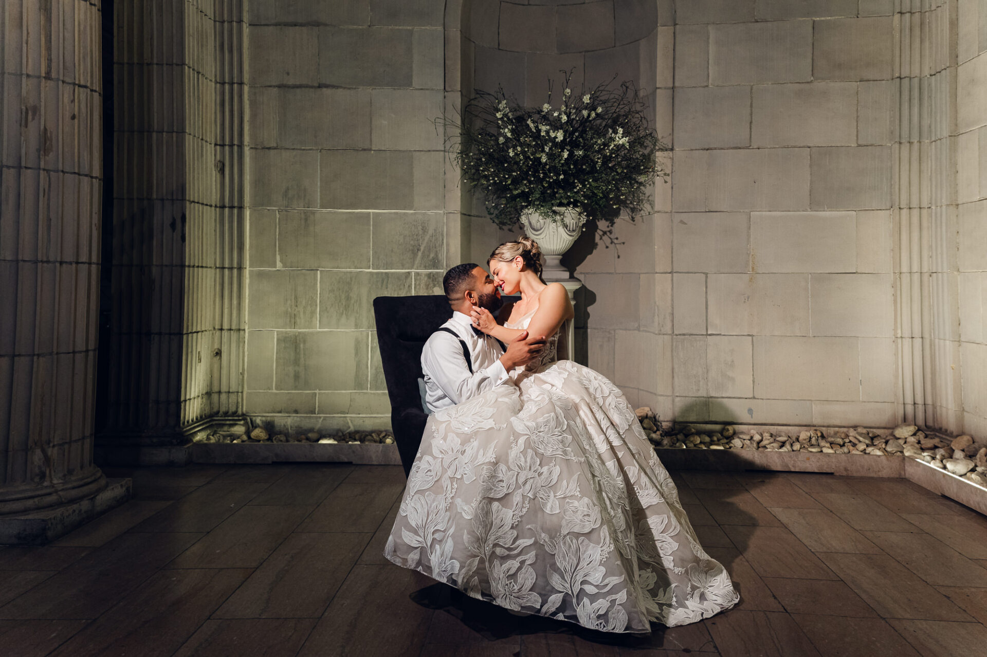 Bride in a floral strapless gown sitting on the groom's lap in a dark armchair, leaning in close with her hand on his jaw inside a stone alcove with a floral arrangement behind them.