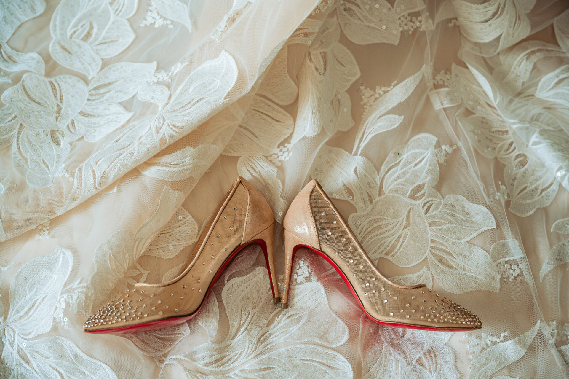 Rhinestone bridal heels with red soles rest on the floral lace fabric of brides wedding gown