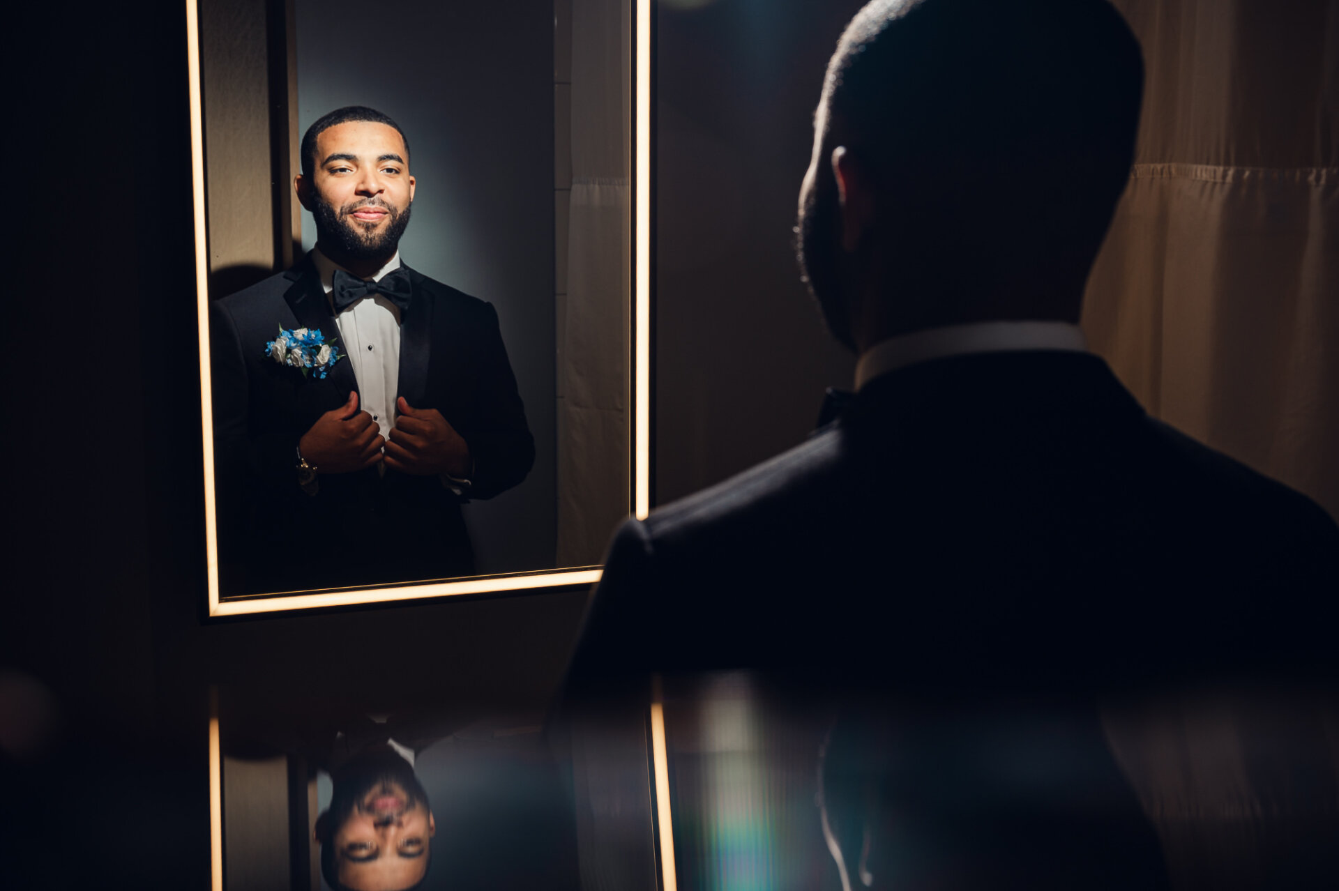 Groom checks his black tuxedo and blue boutonniere in an illuminated mirror.