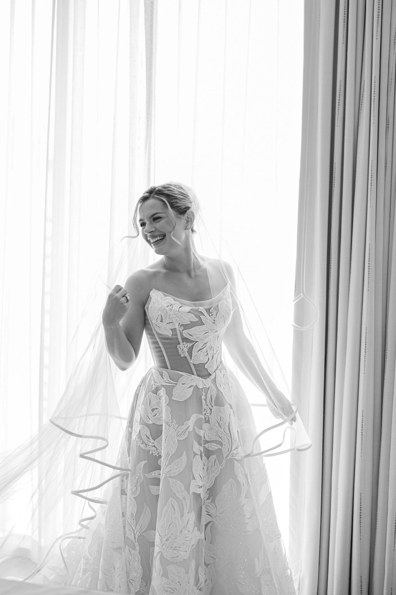 bride smiles beside a bright window in her floral lace gown and veil before her wedding at The Ben Philadelphia