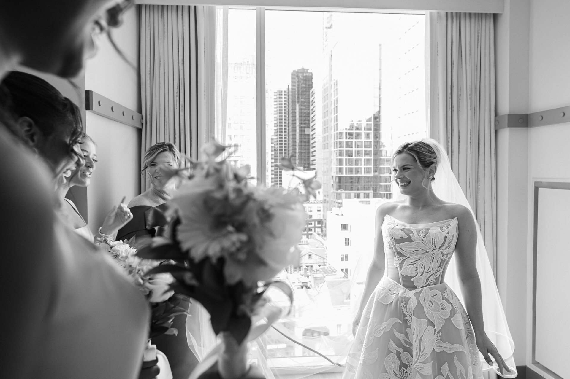 Bride smiles with her bridesmaids beside a window before the wedding ceremony.