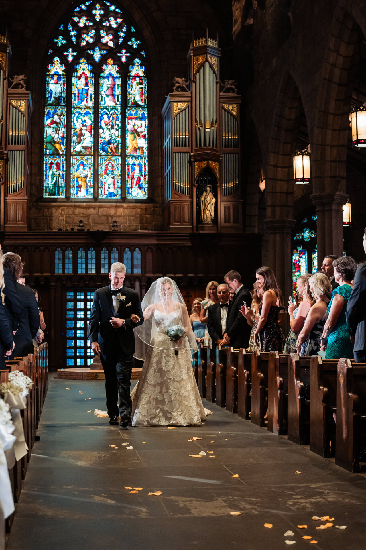 Bride walks down the aisle with her father beneath colorful stained-glass windows.