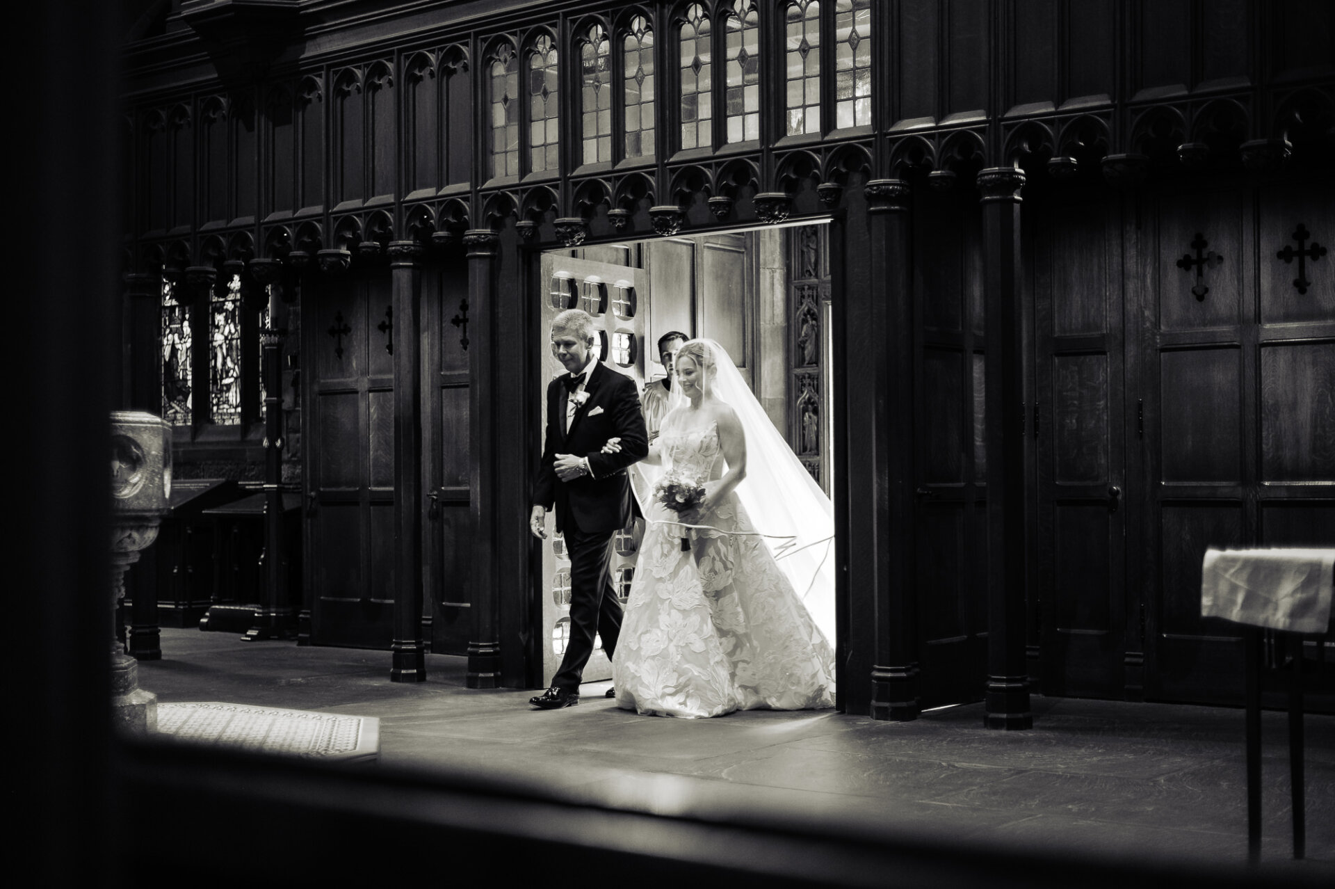 Bride walks down the church aisle with her father in a black-and-white wedding photo.