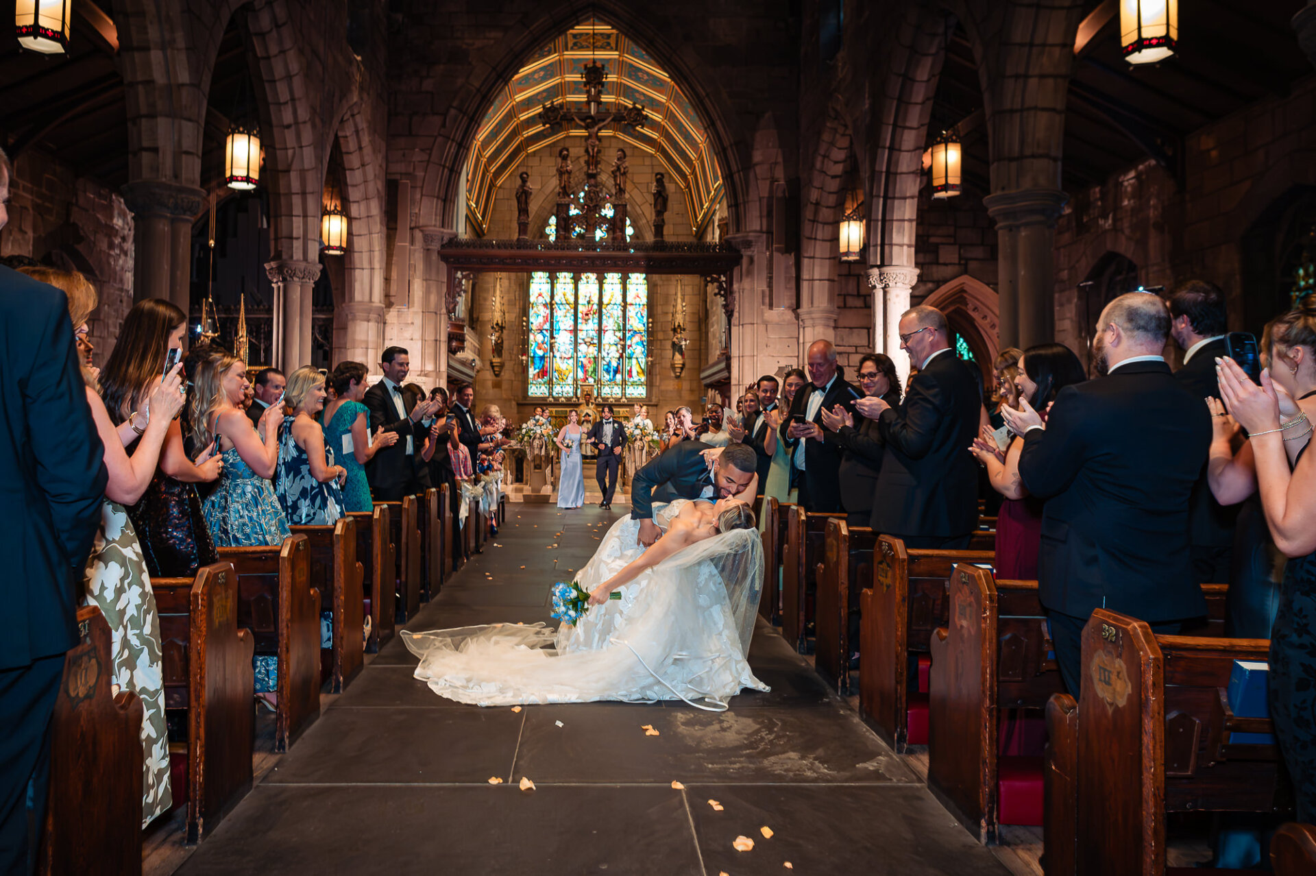 Groom dips and kisses the bride in the aisle as guests celebrate their church wedding ceremony.