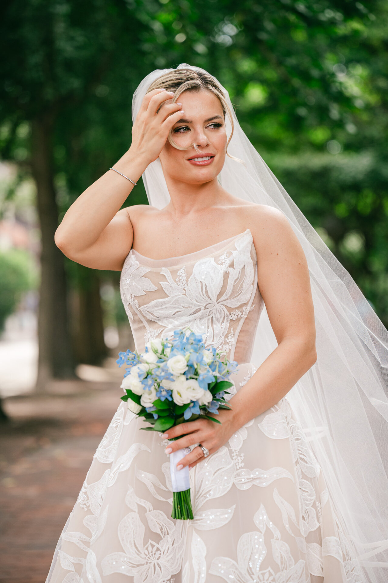Portrait of a bride holding her white and light blue floral bouquet while touching her hair outdoors, wearing a sheer corset-style gown and veil.