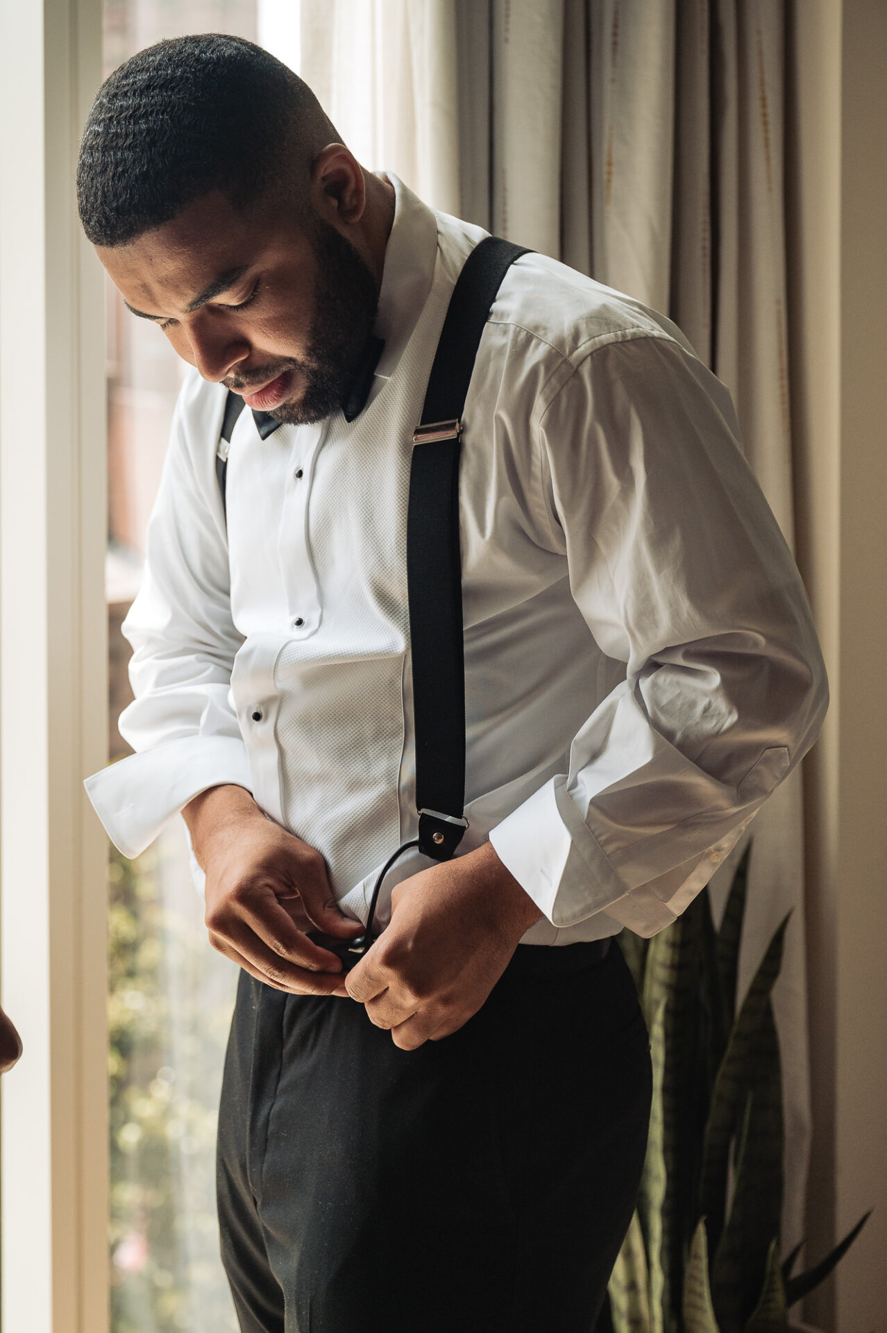 groom fastens his black suspenders while getting ready in a white dress shirt and bow tie.